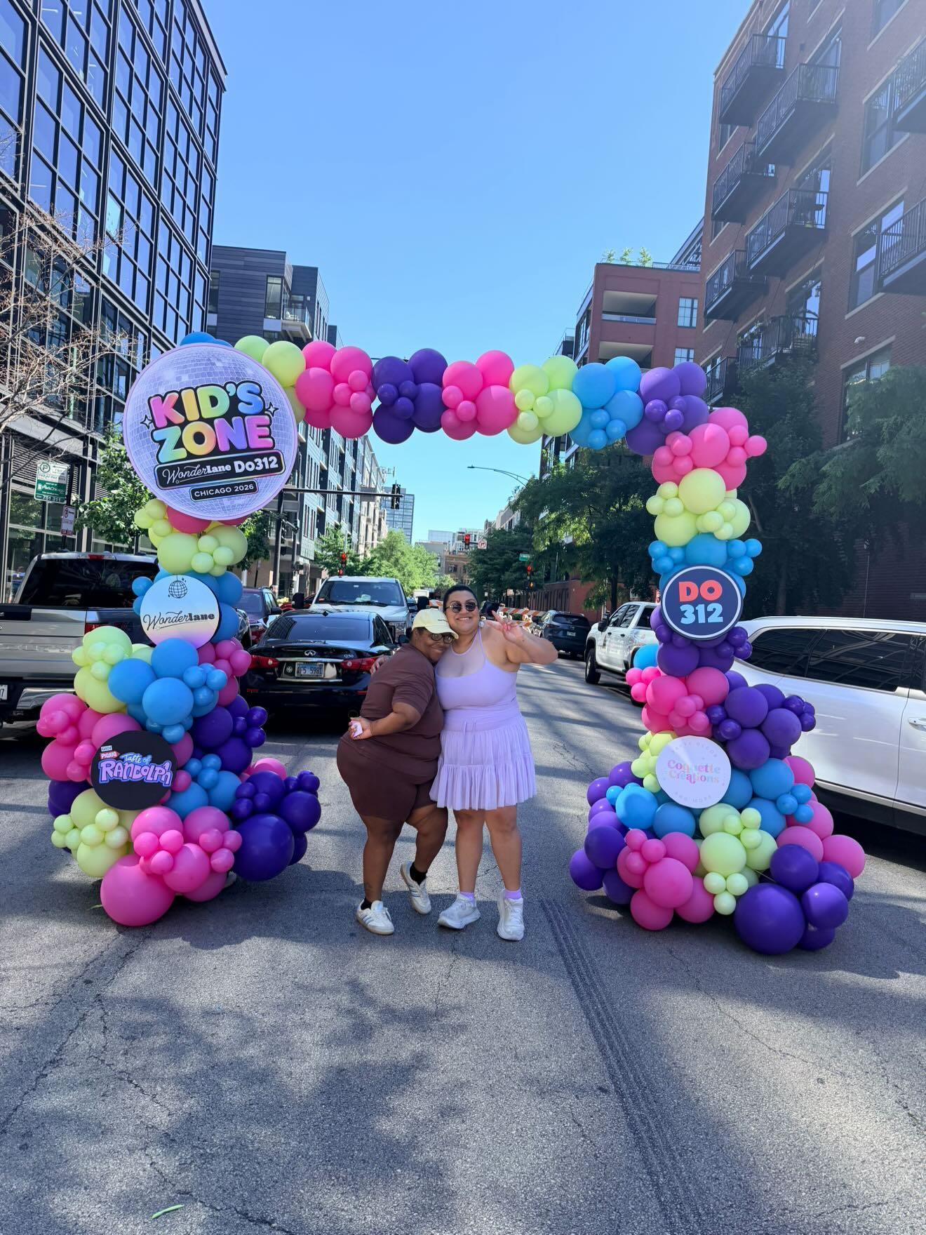 Two children stand under colorful balloon arch on city street, with buildings and cars in background, sunny day.