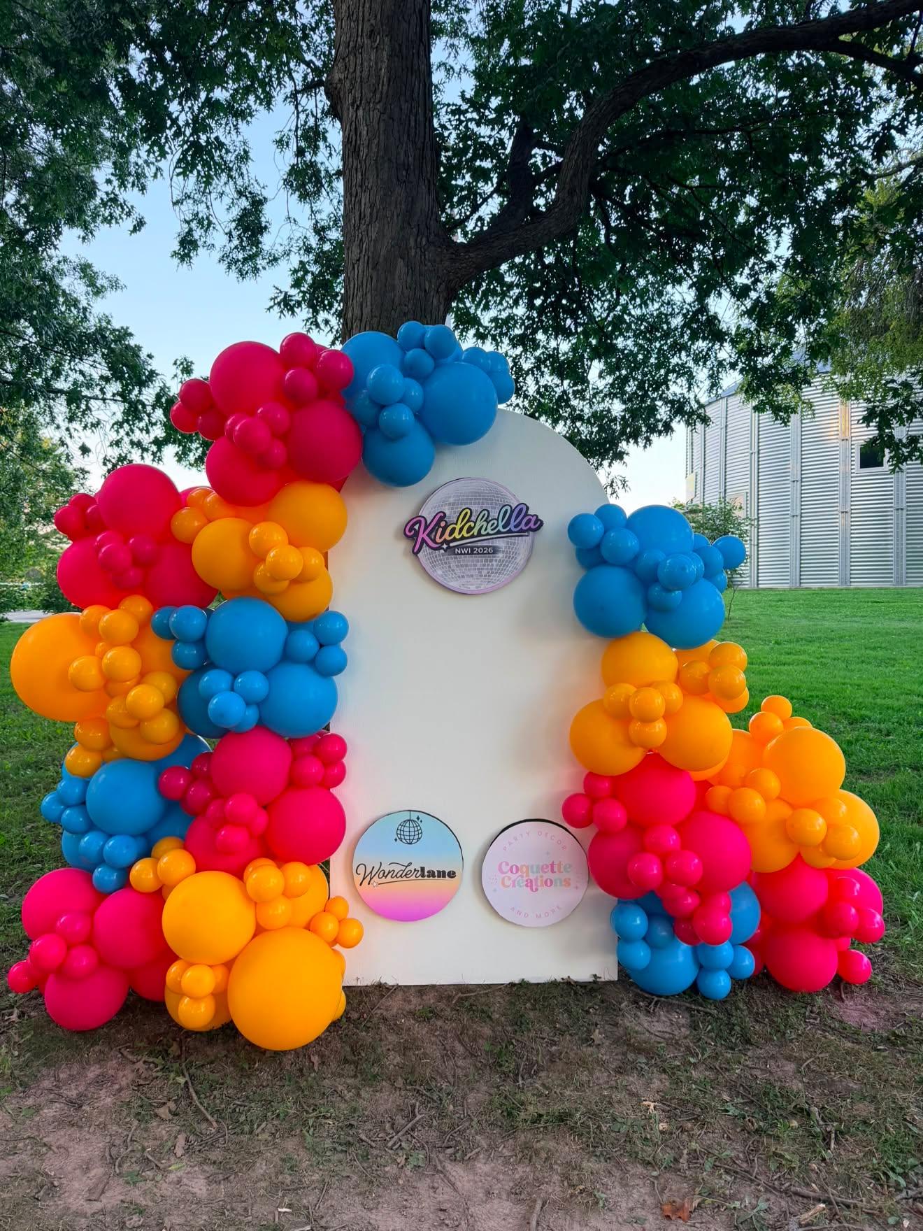 Decorative display with colorful balloons in red, yellow, blue, and pink around a white backdrop, outdoors under a tree.