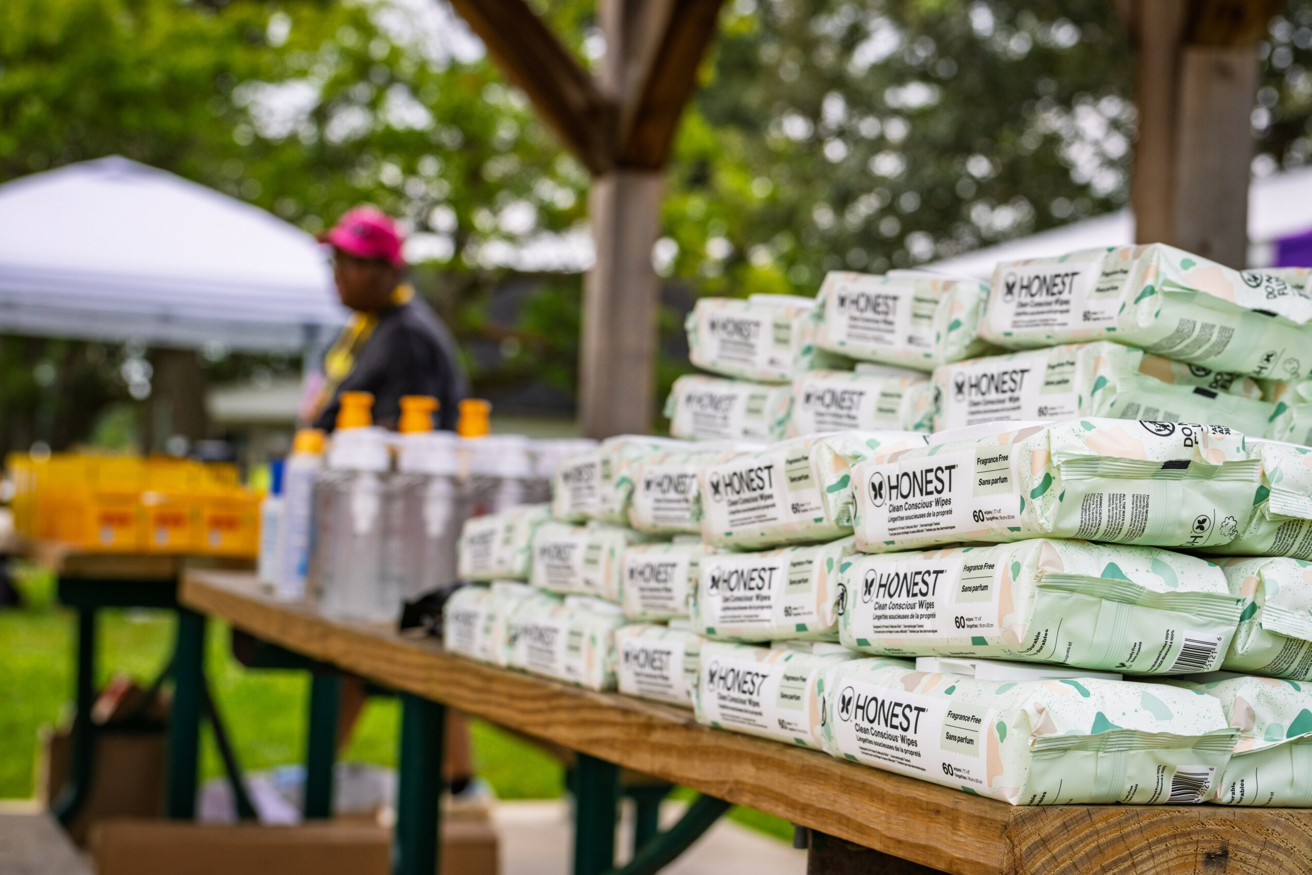 Stacks of packaged wipes on a wooden table outdoors, with a person in the background near a tent and trees.