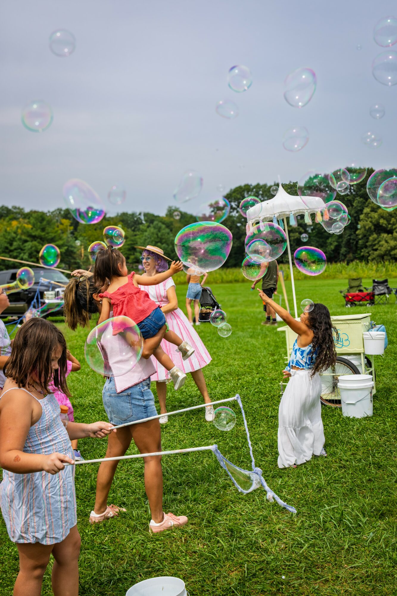 Children and adults playing with bubbles outdoors on a grassy field, with trees and a cloudy sky in the background.