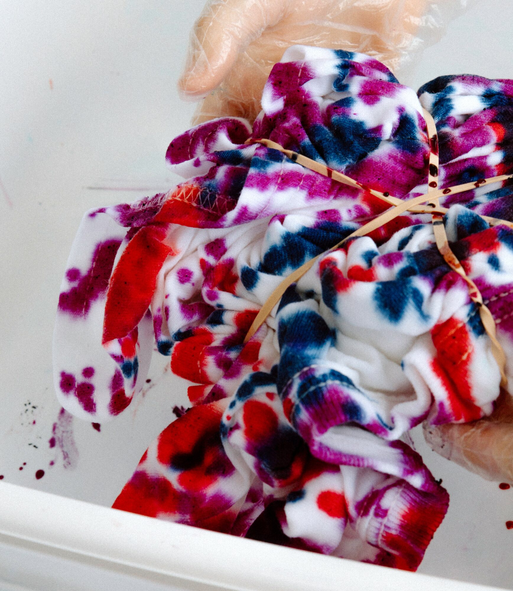 Person holding a tie-dye fabric bundle with a rubber band, colorful red, blue, purple, and white pattern, in a white basin.