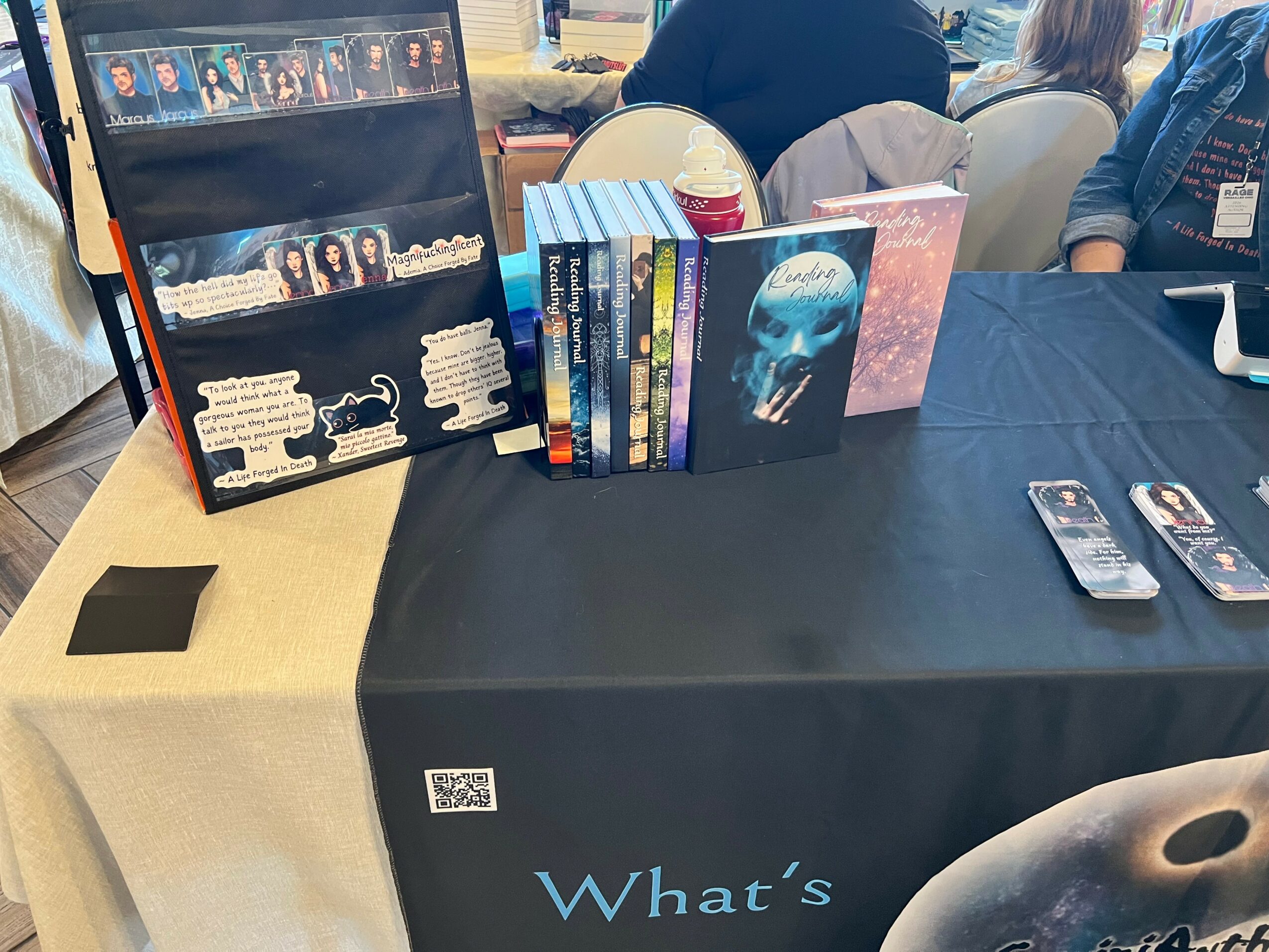 Table with books and merchandise, including a display stand and a box of books, at a convention or event.
