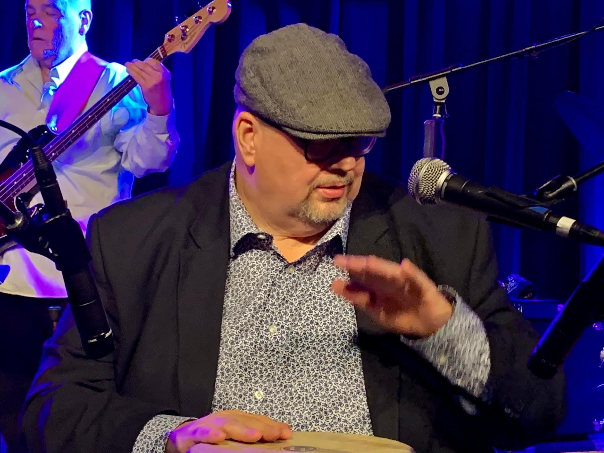 Man wearing a gray cap, glasses, and patterned shirt, speaking into a microphone on stage with a musician in the background.