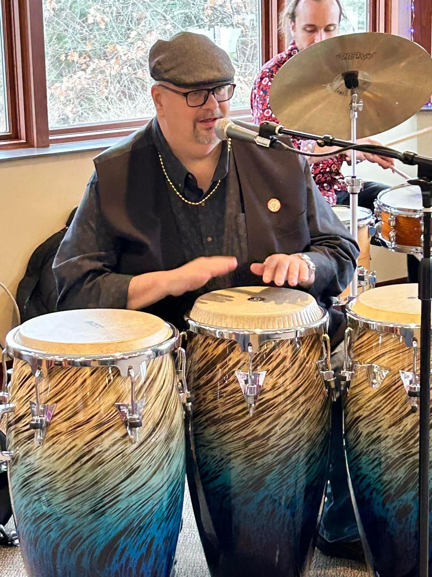 Person wearing glasses and a hat playing conga drums with a microphone nearby, indoors with windows in the background.