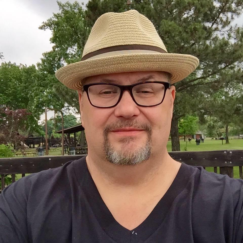 Man wearing glasses and a straw hat outdoors, smiling, with trees and a park in the background.