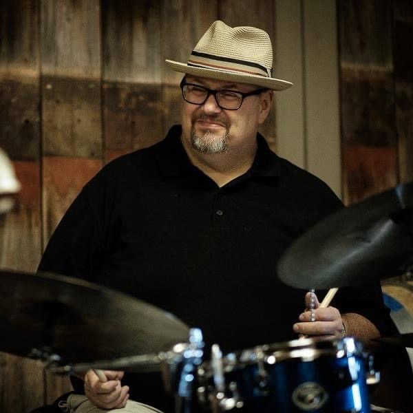 Man wearing a hat and glasses playing drums, wooden background, holding drumsticks, smiling.