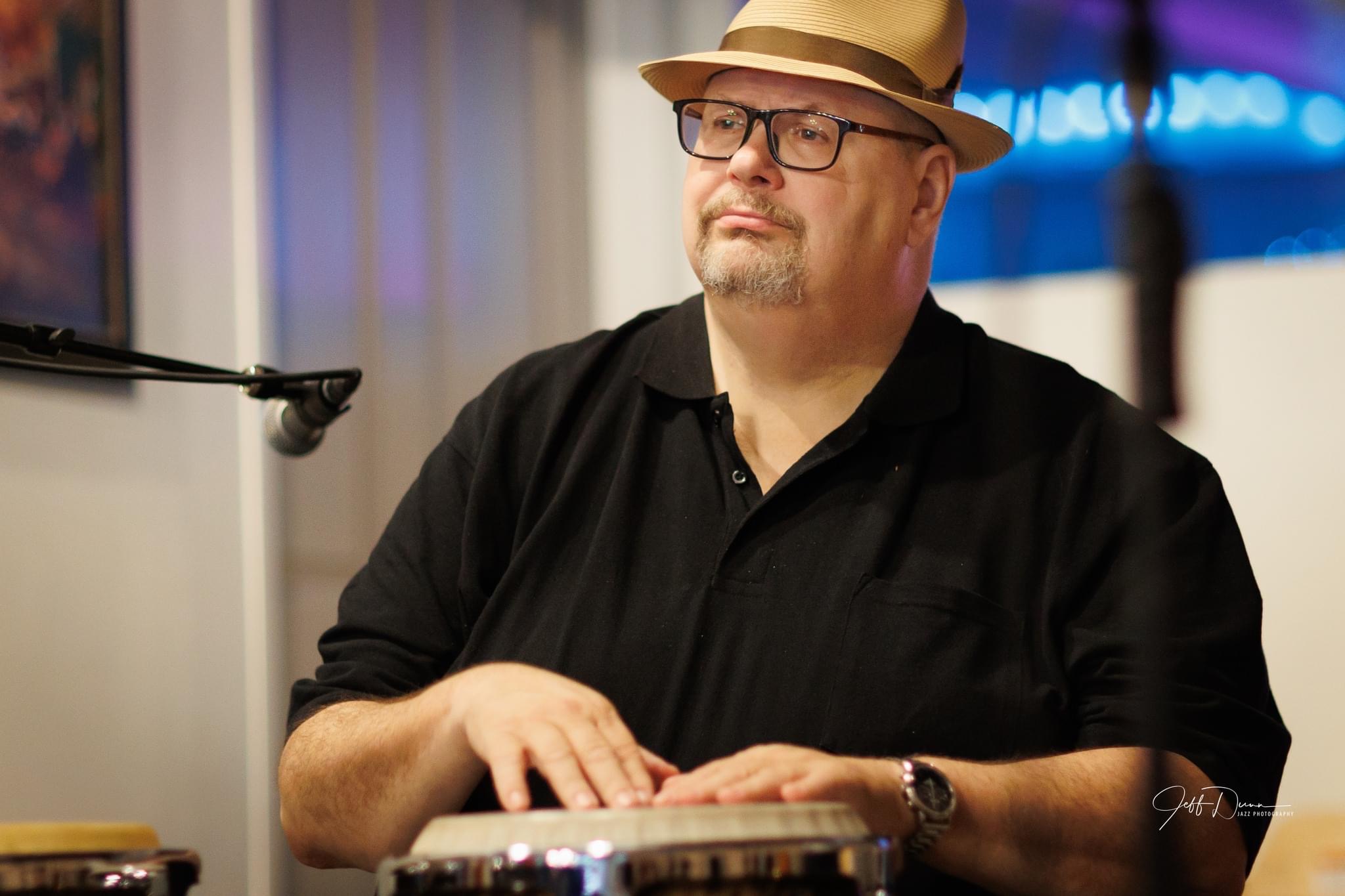 Man wearing glasses, a hat, and a black shirt playing a keyboard indoors, with a microphone nearby and colorful background.