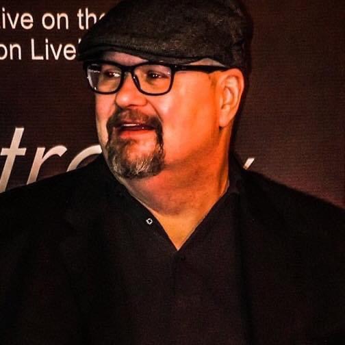 Man with glasses, beard, and mustache wearing a black cap and black shirt, smiling, against a dark background.
