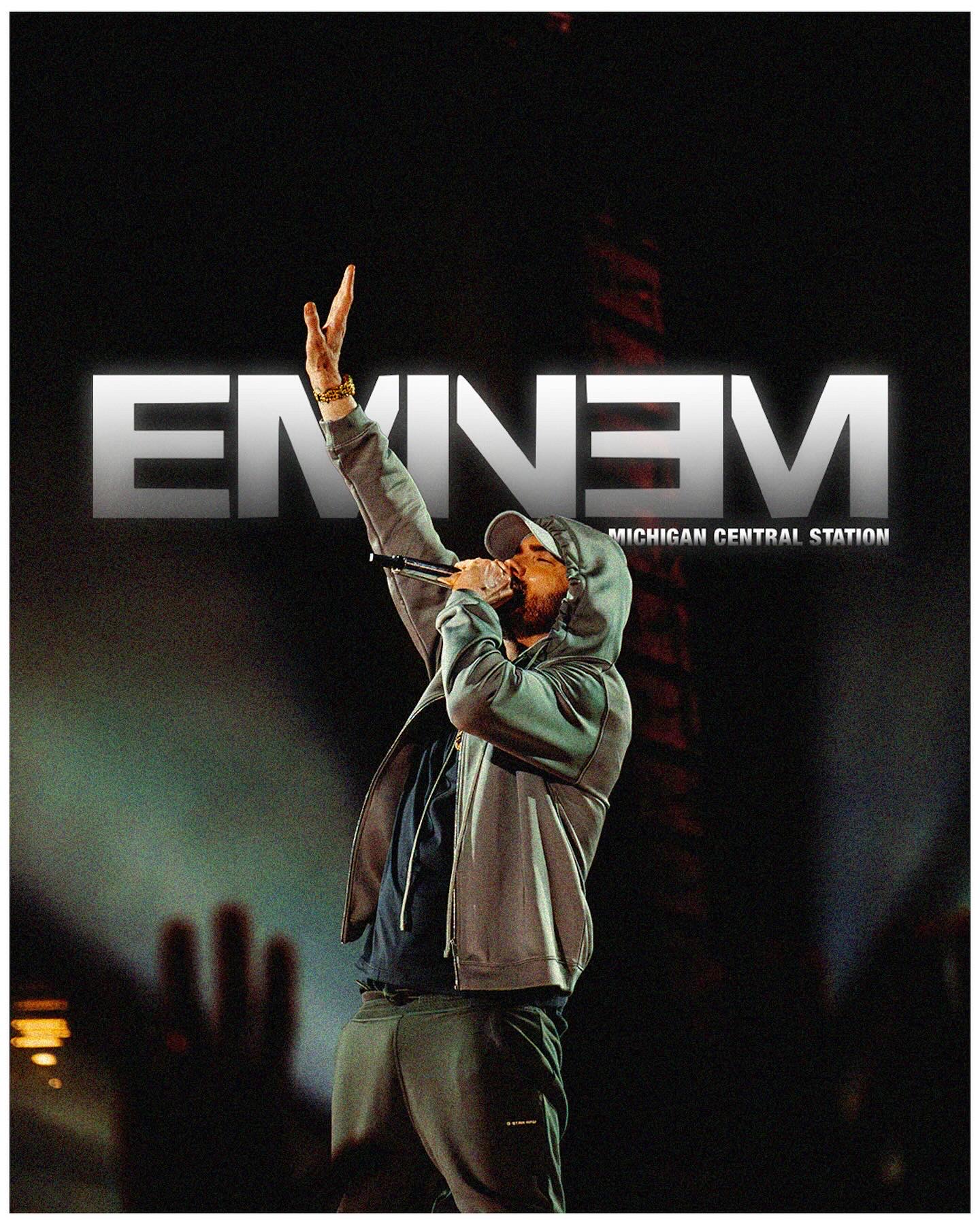 Performer on stage with microphone, hand raised, background shows large illuminated text, audience in foreground.