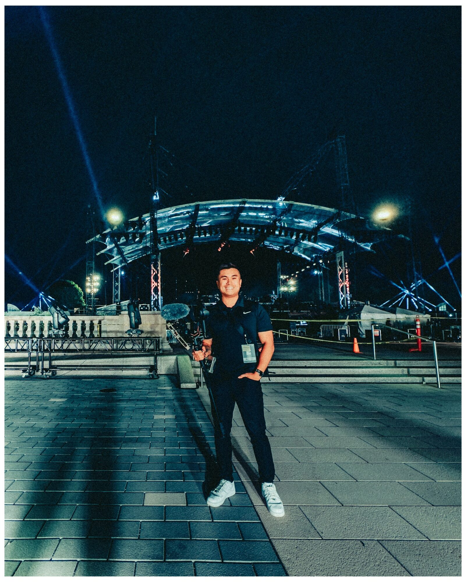 Person standing outdoors at night with illuminated stage and structures in background.