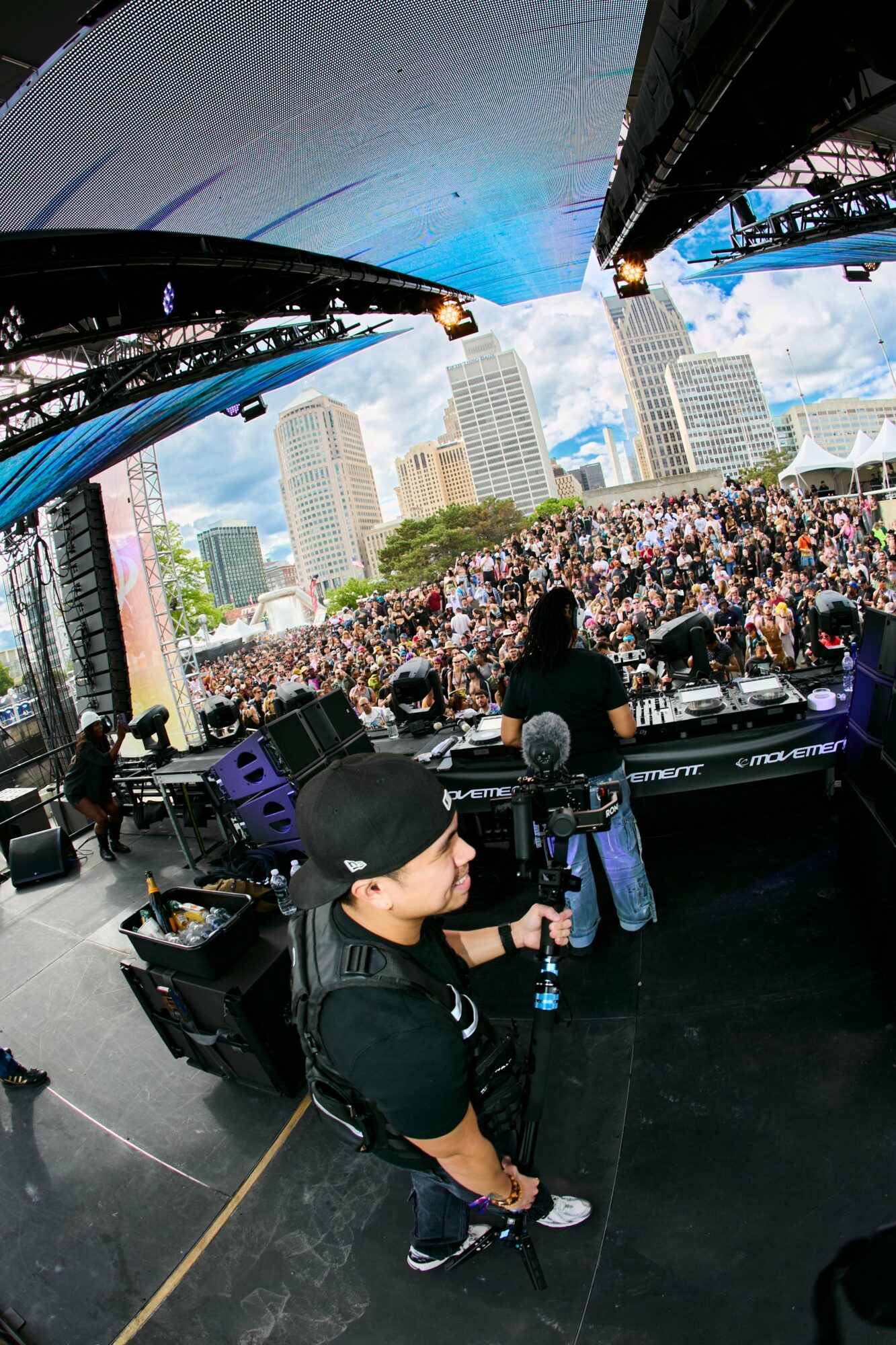 DJ performing on stage with a large crowd and city skyline in background, stage overhead with blue sky and clouds