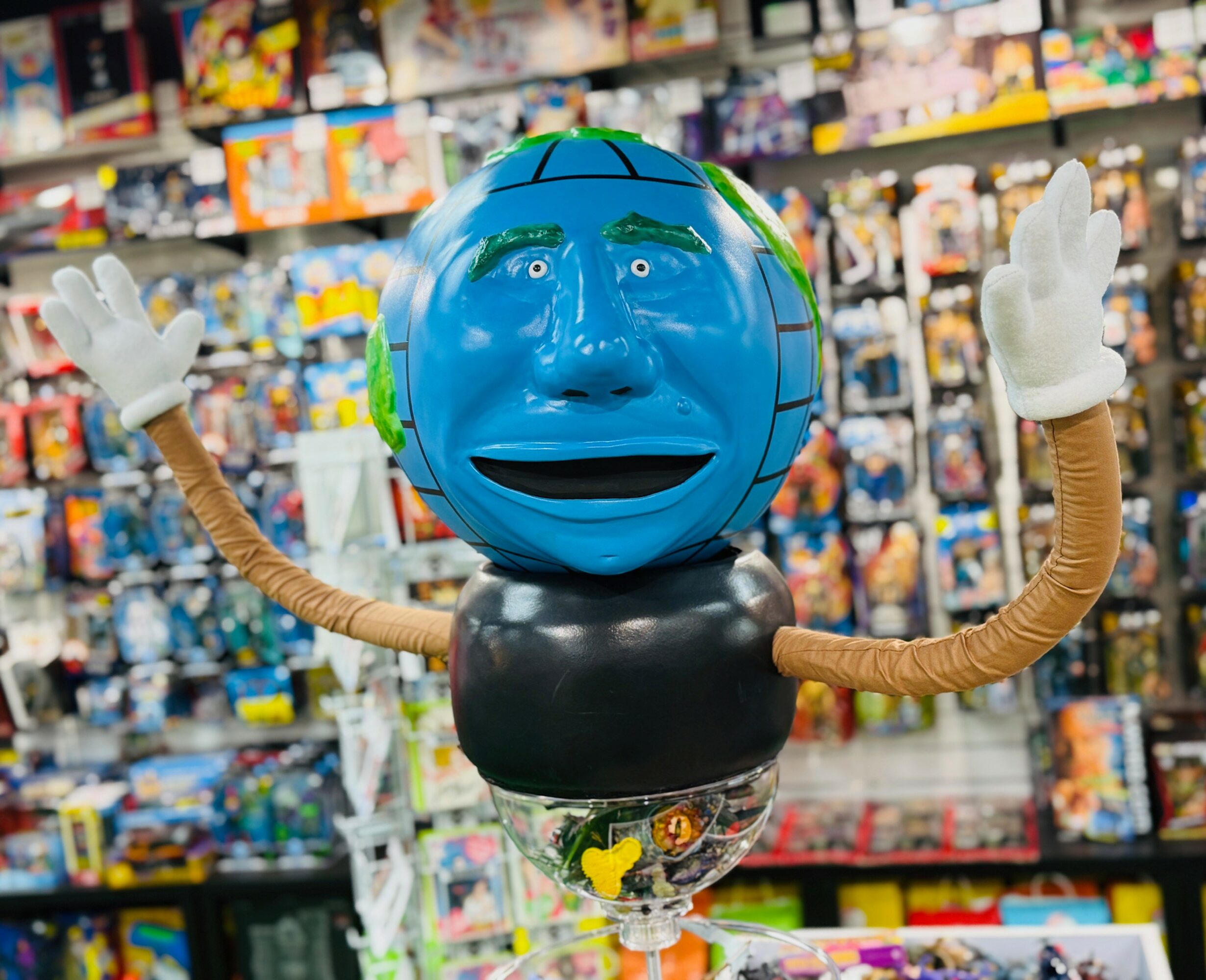 A balloon figure with a blue face, black body, and arms made of twisted material, in a store aisle filled with toys.
