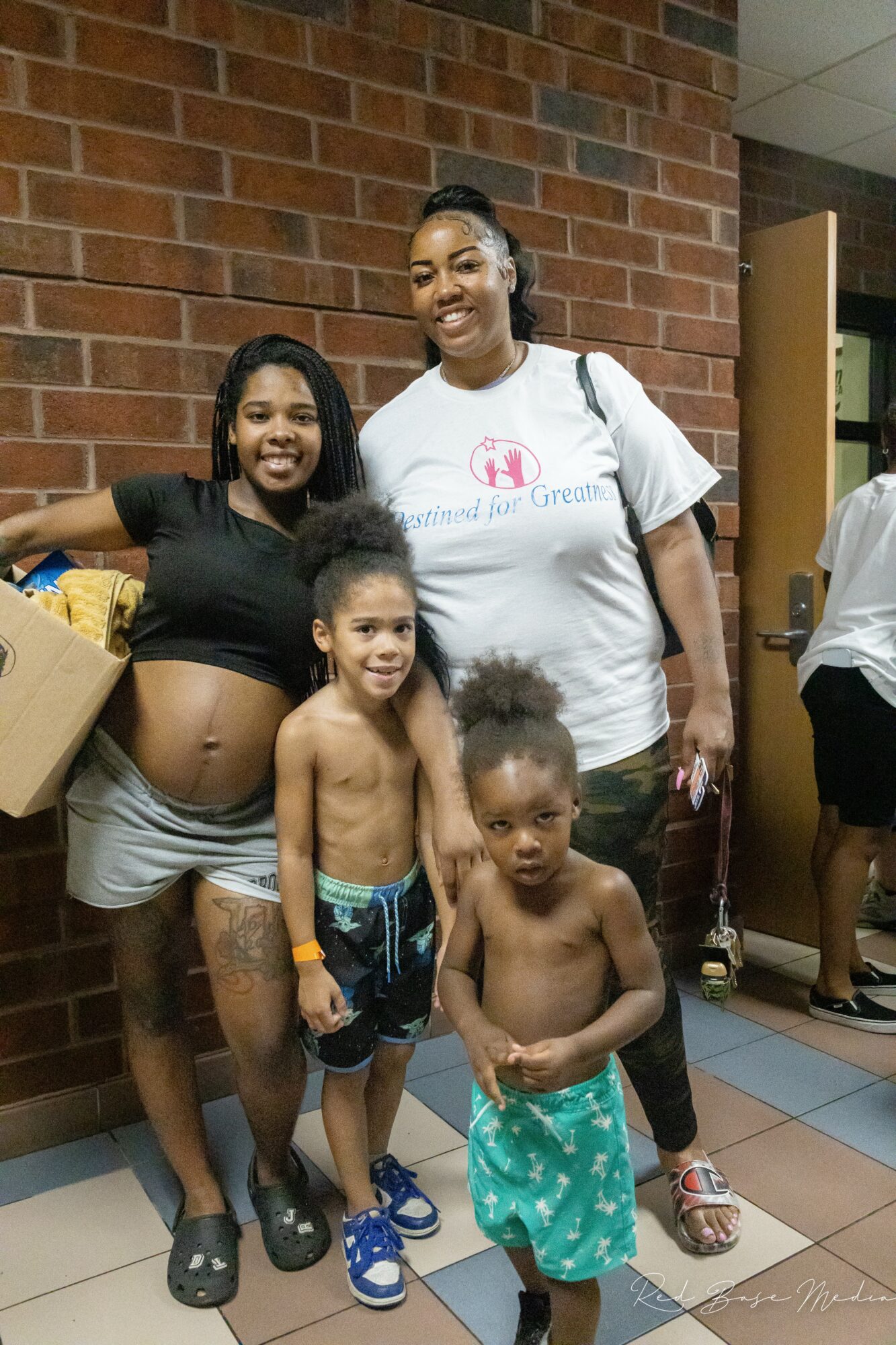 Four people, including two children, stand together indoors in front of a brick wall, smiling at the camera.