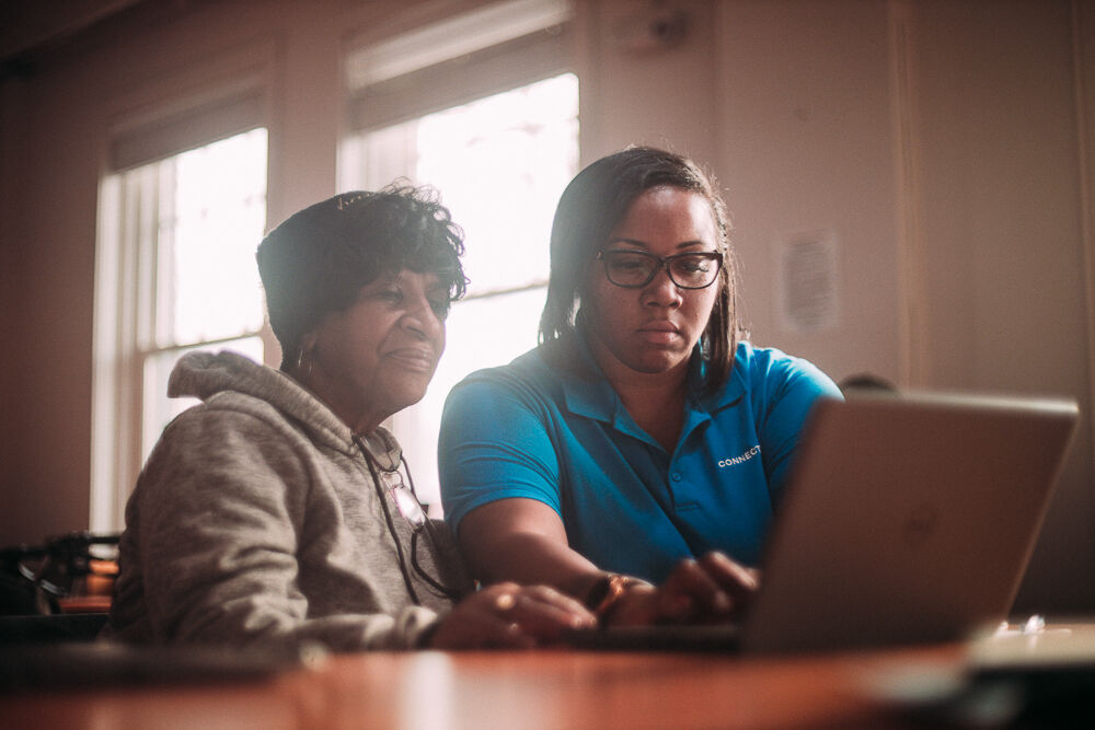 Two women sit at a table looking at a laptop screen, with windows in the background. One woman wears glasses and a blue shirt, the other wears a hoodie.