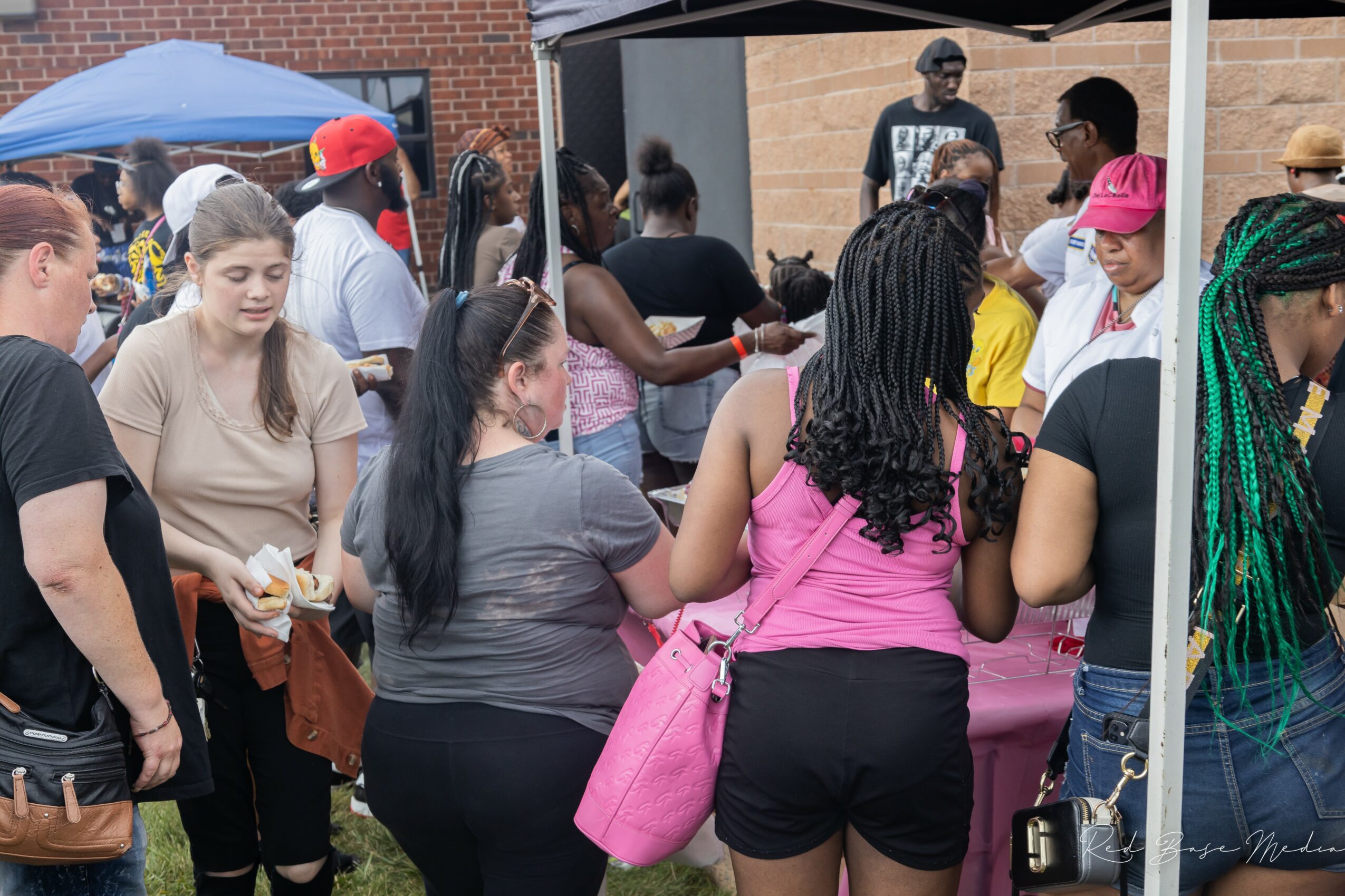 Group of people gathered outdoors around a table, some holding food, with a brick building in the background.