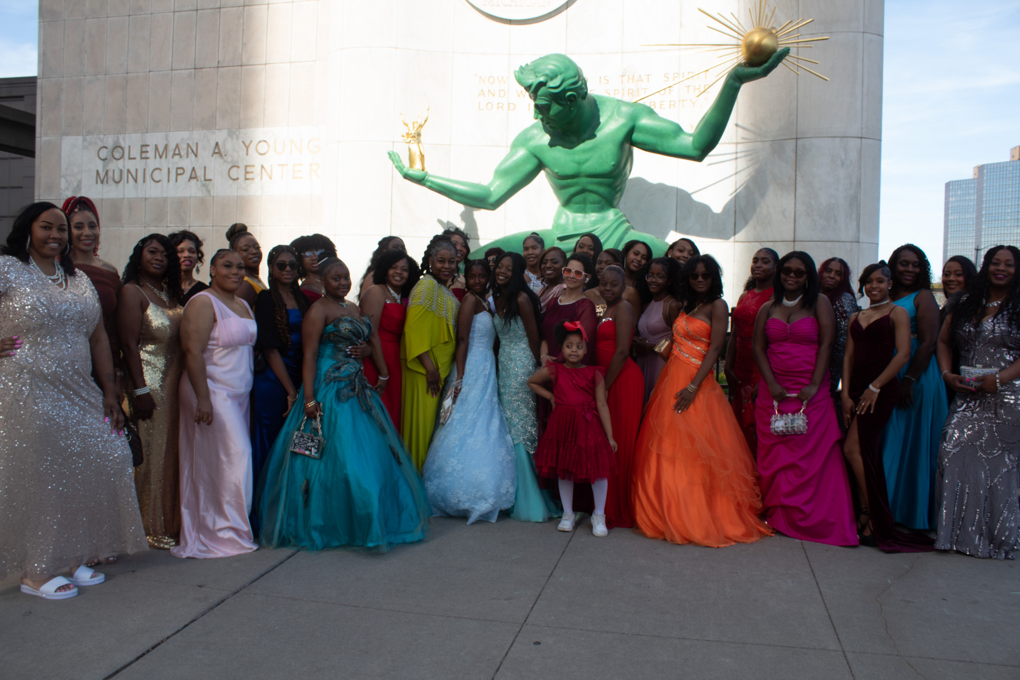 Group of women in colorful formal dresses standing in front of a large green statue outside a municipal center.