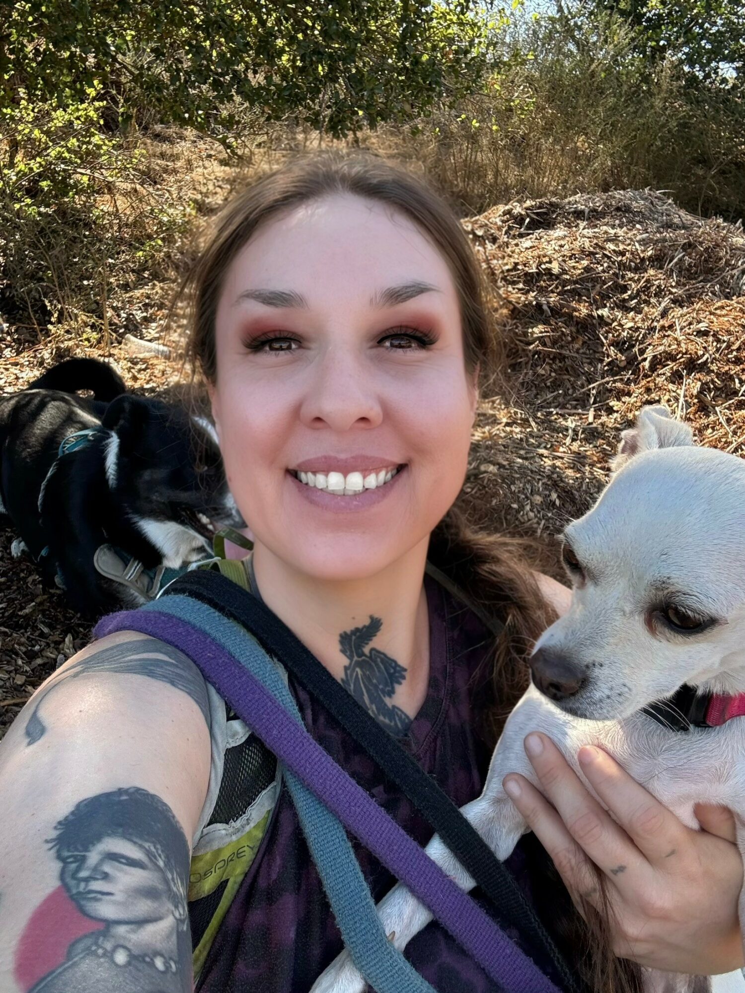 Smiling woman holding a white dog outdoors with trees and dry grass in background.