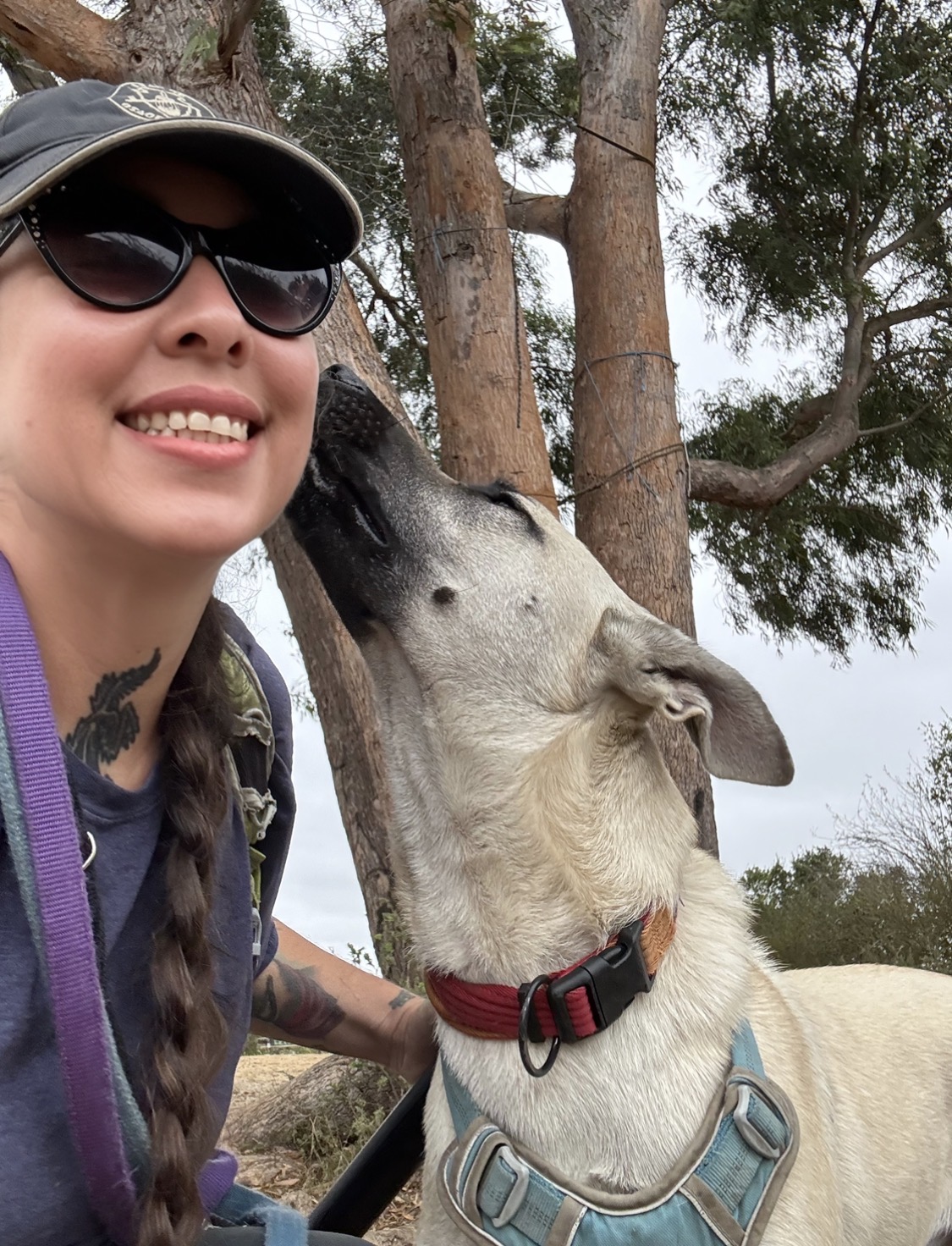 Person smiling with sunglasses and a hat, close to a dog outdoors near trees.