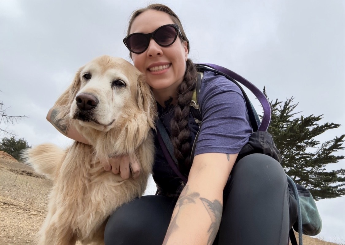 Woman with sunglasses smiling next to a golden retriever outdoors, cloudy sky and trees in background.
