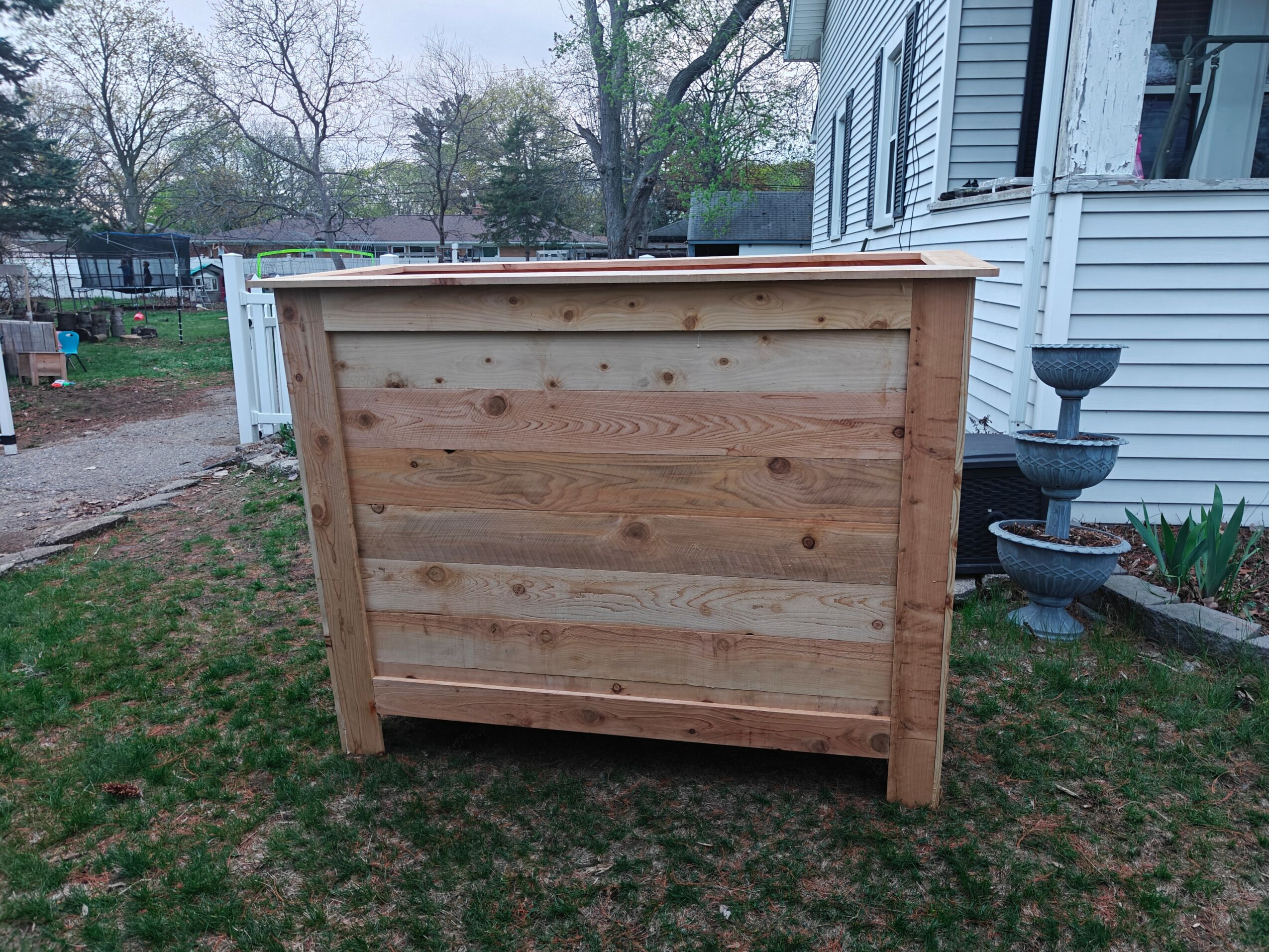 Wooden outdoor storage box on grass in front of a house with trees in background.