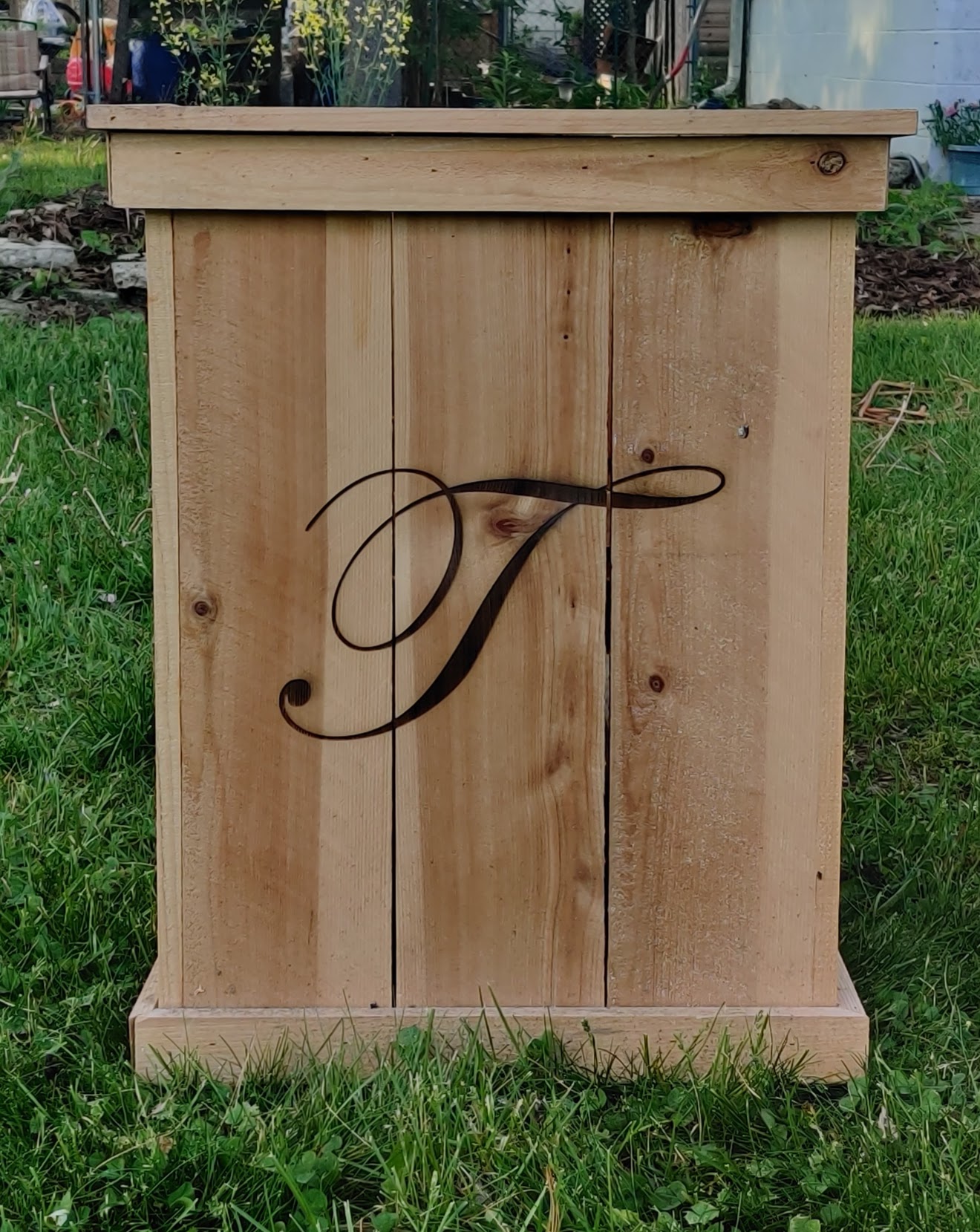 Wooden cabinet with a decorative black letter 'J' on the front, placed outdoors on grass.