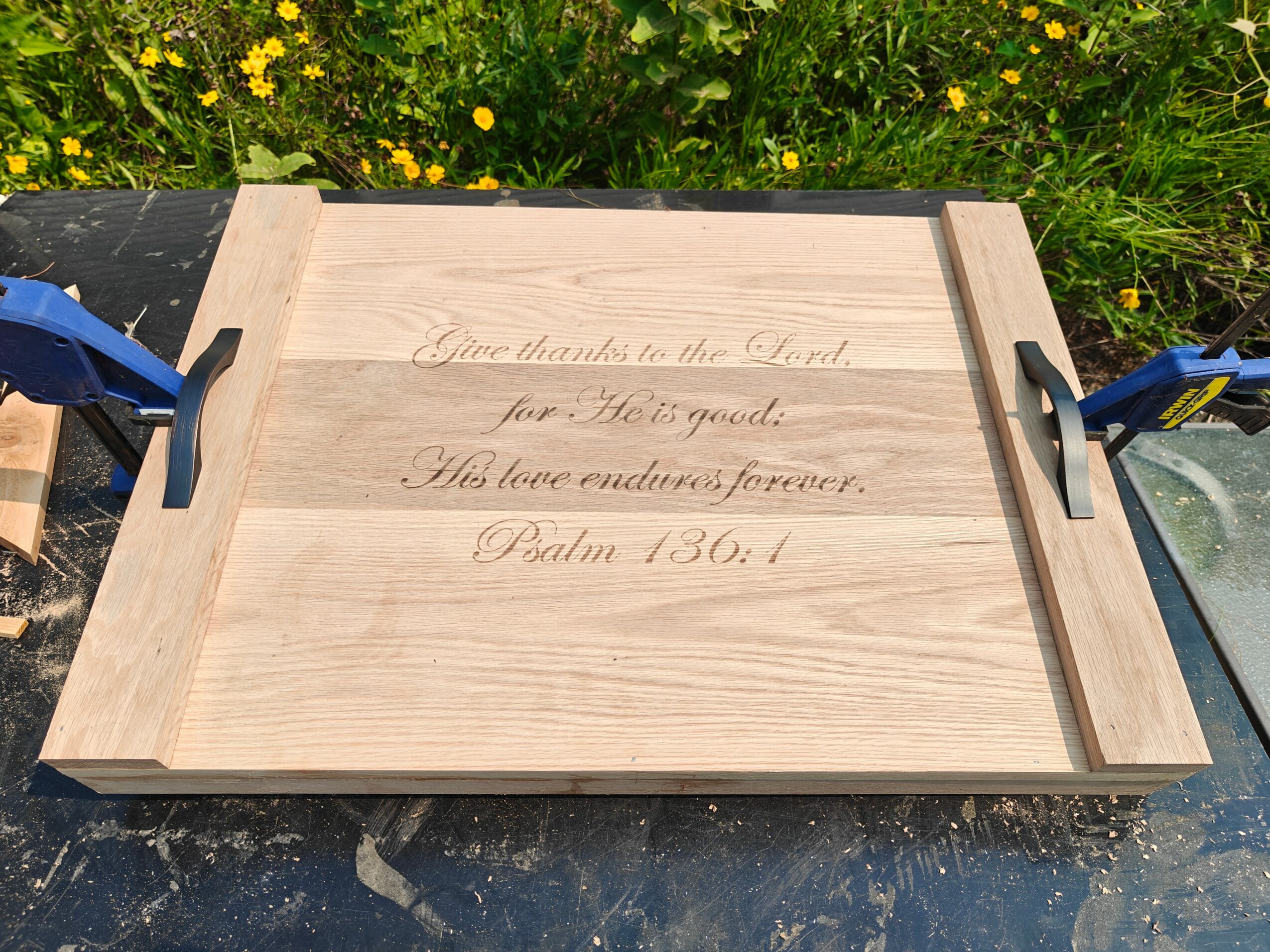 Wooden tray with engraved text, held by clamps outdoors with yellow flowers in background.