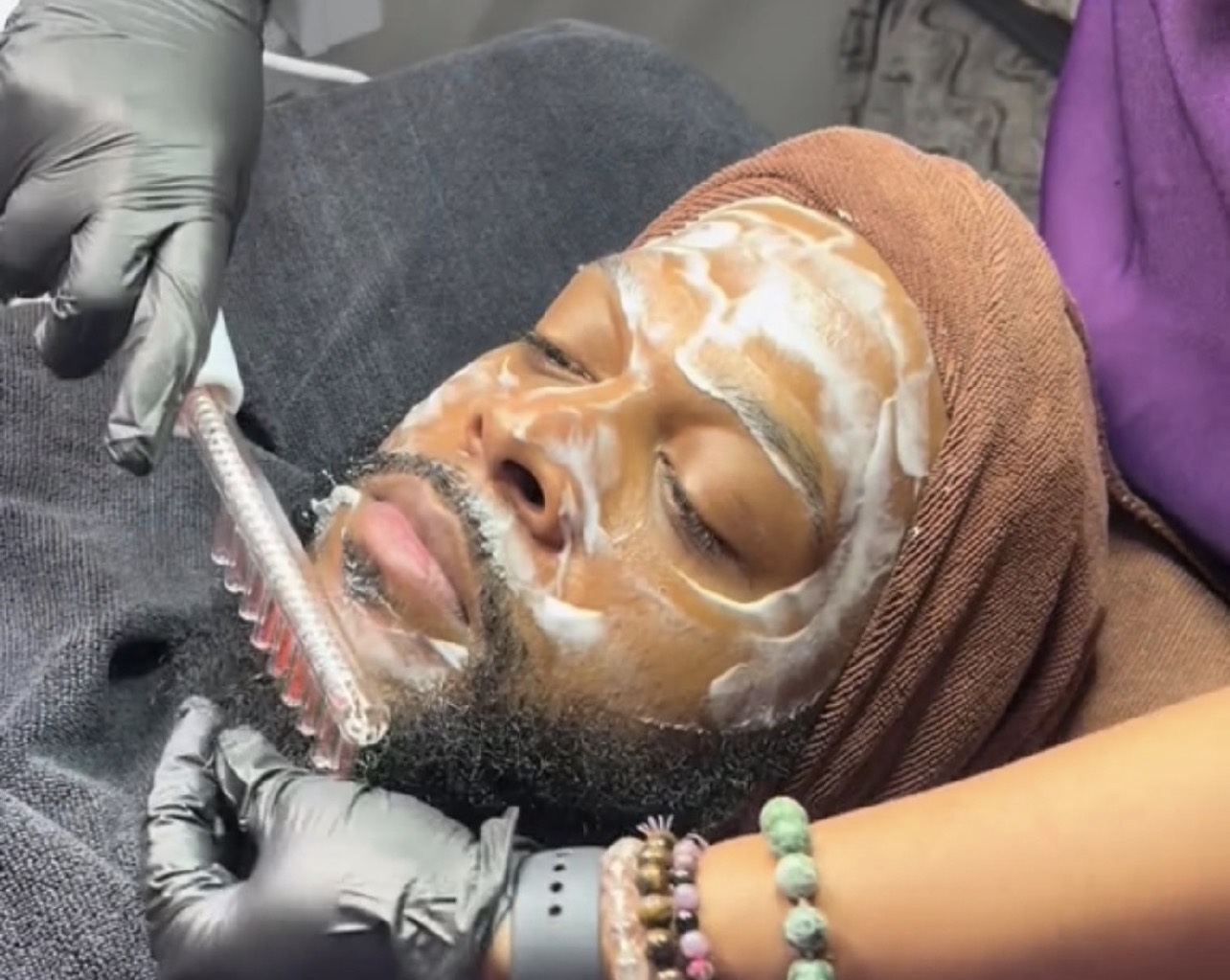 Person lying down with a facial mask, surrounded by medical or skincare professionals wearing gloves, performing a treatment.