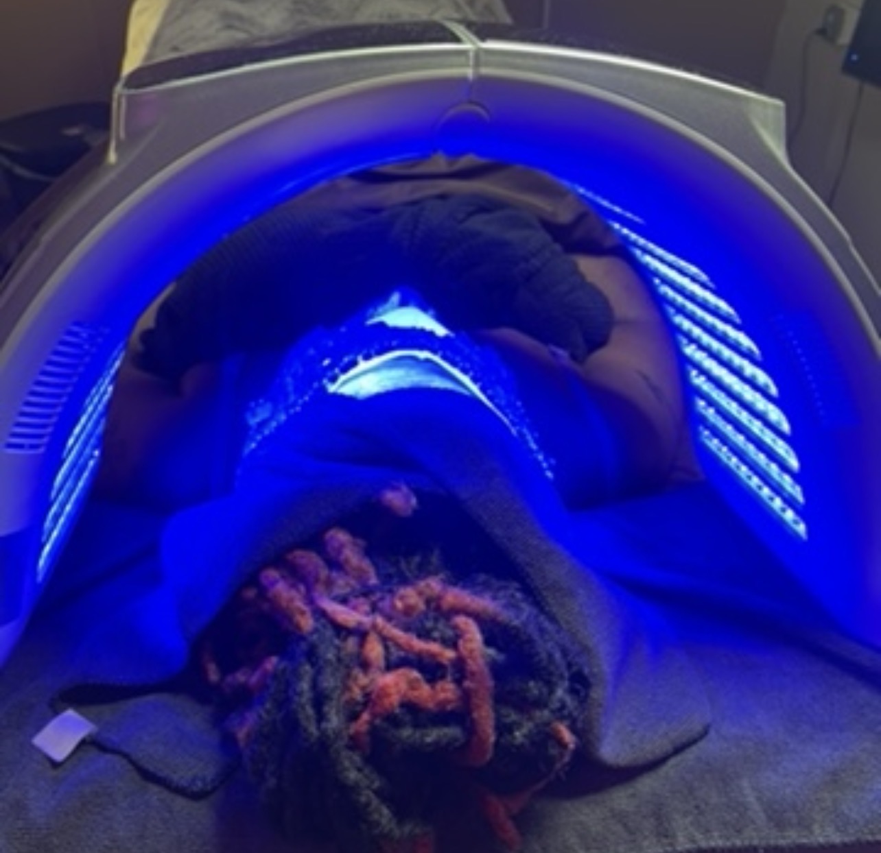 Person lying inside a blue-lit medical or tanning device, head facing downward, with dreadlocks visible. Word count: 20