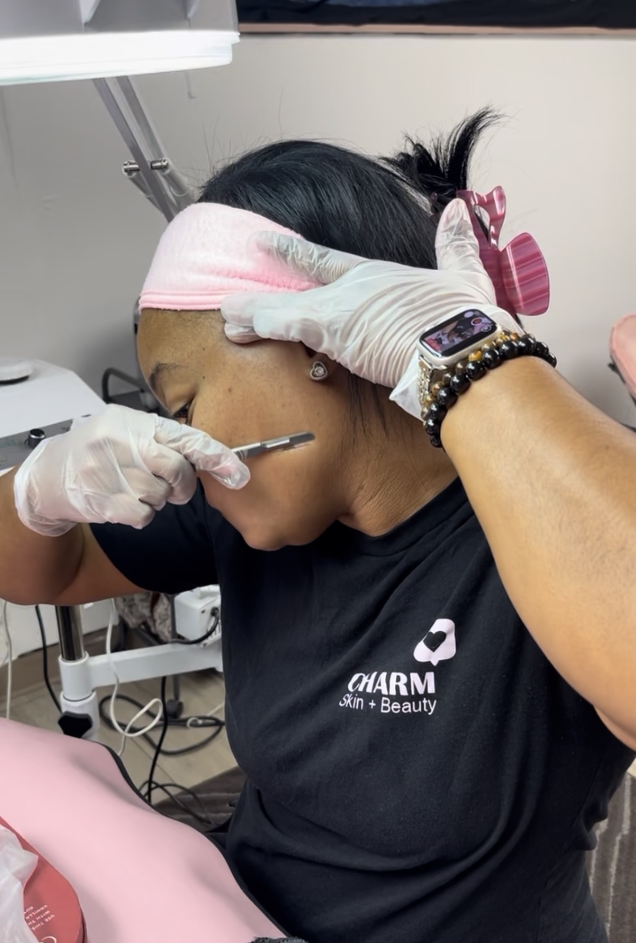 A woman receives a cosmetic injection in a clinical setting, wearing gloves and a headband, with medical equipment nearby.