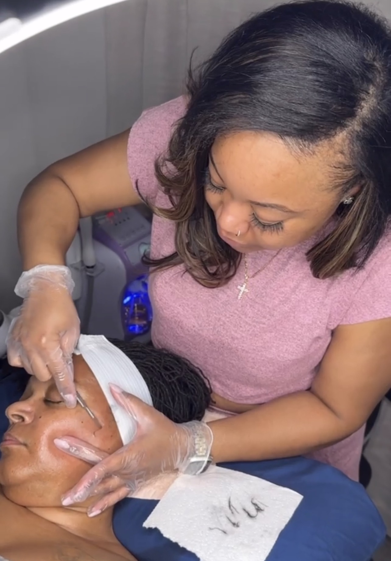 Woman performing a facial treatment on a person lying down, with a towel covering their face, in a clinical setting.