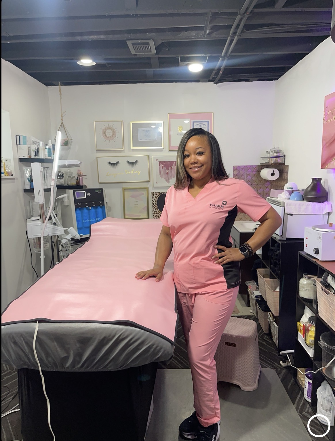 Smiling woman in pink scrubs standing in a medical or spa room with treatment bed and equipment, white walls, and colorful artwork.
