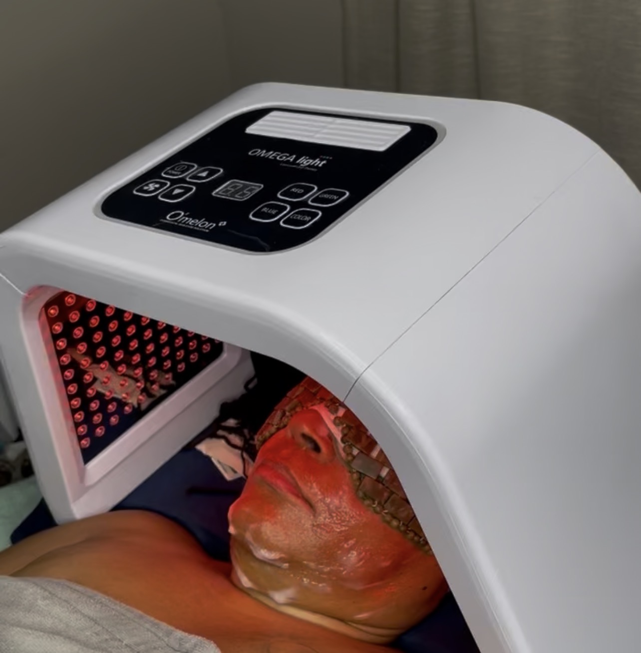 A person receiving a facial treatment under a white LED light device, with their face covered in a mask.