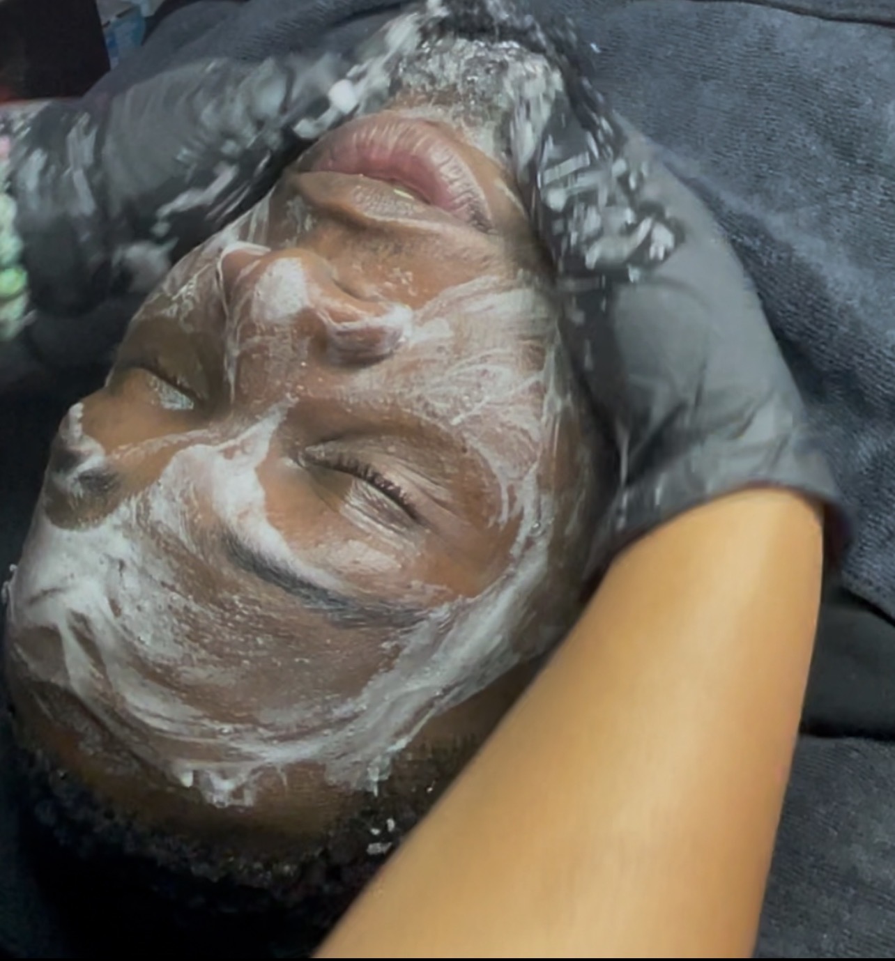 Person receiving facial treatment with cream applied, lying down with eyes closed, surrounded by hands and towels.