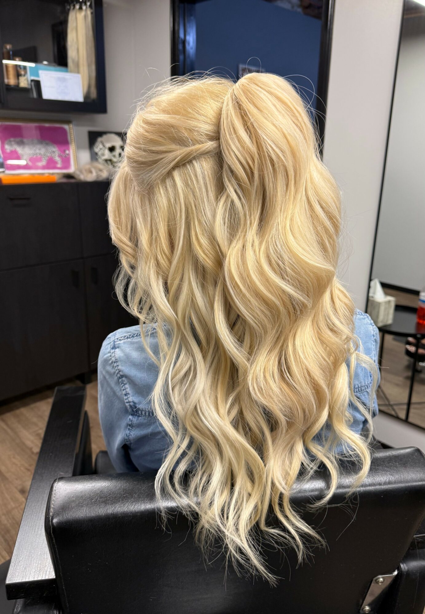 Back view of a woman with long, wavy blonde hair in a salon chair, wearing a denim jacket.