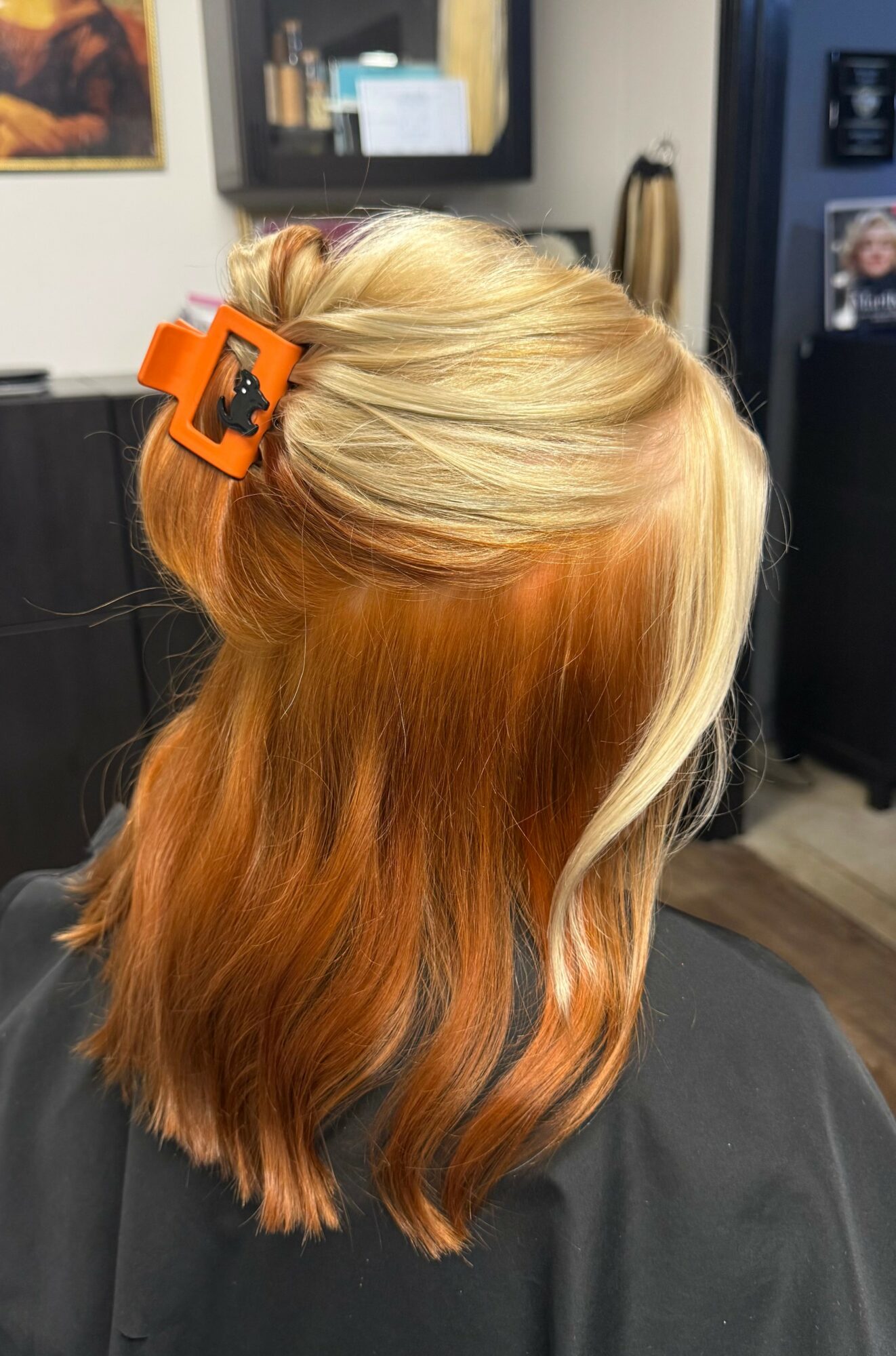 Back of a person's head with blonde and orange hair, held with an orange hair clip, in a salon setting.