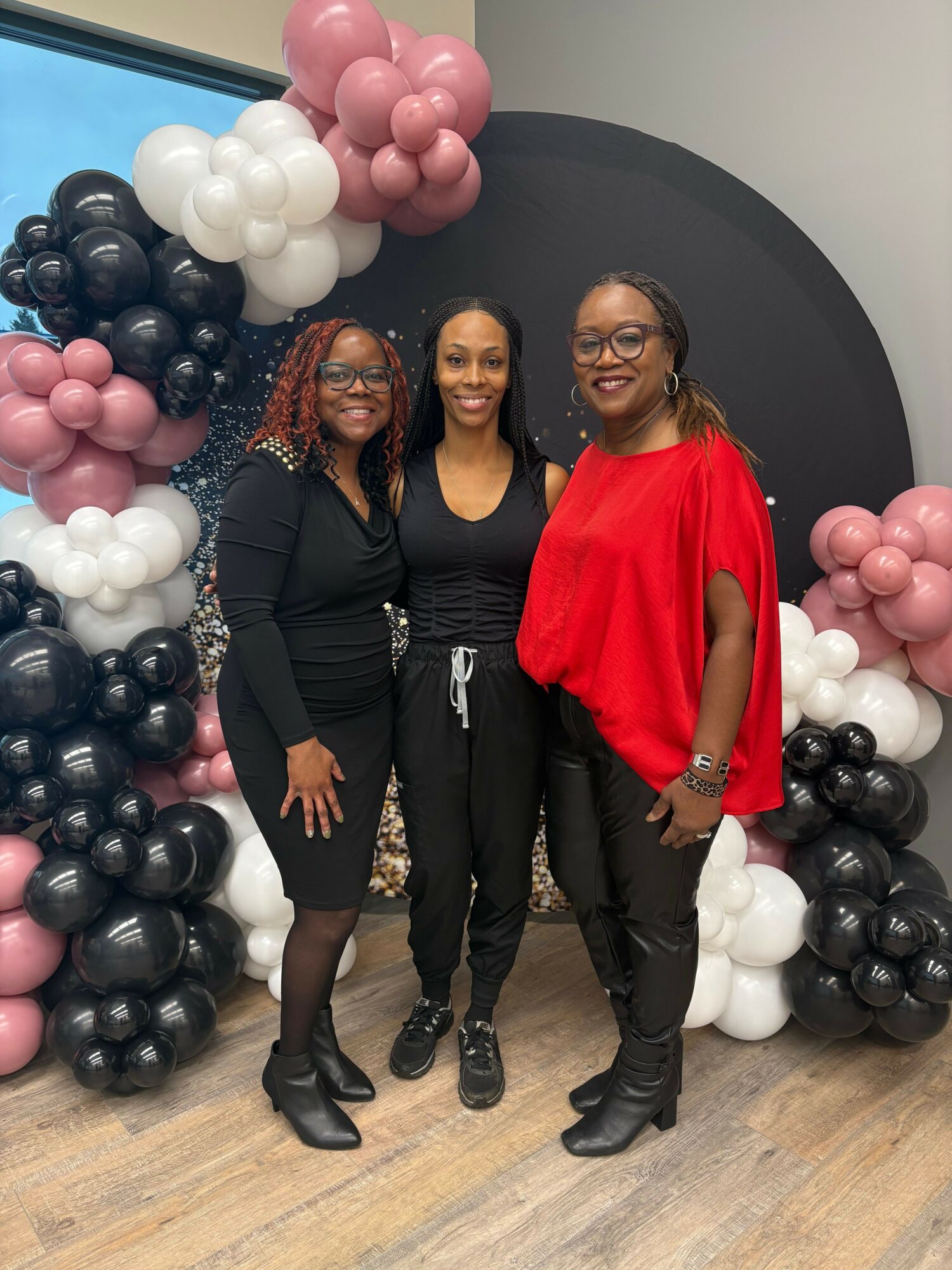 Three women standing together in front of balloon decorations, smiling, with a dark background and wooden floor.