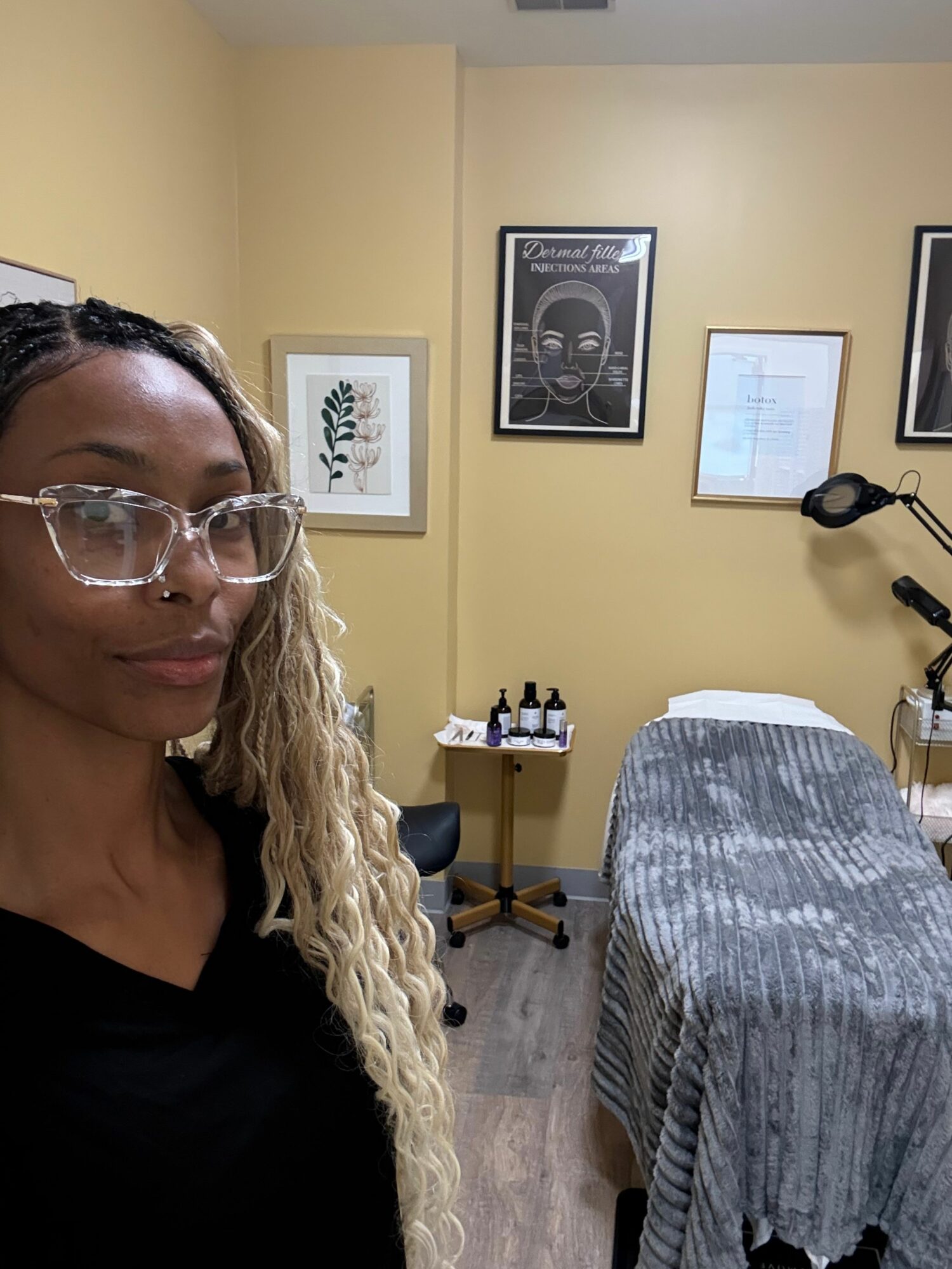 Woman with glasses and long hair taking a selfie in a room with framed pictures on the wall and a bed with a striped blanket.
