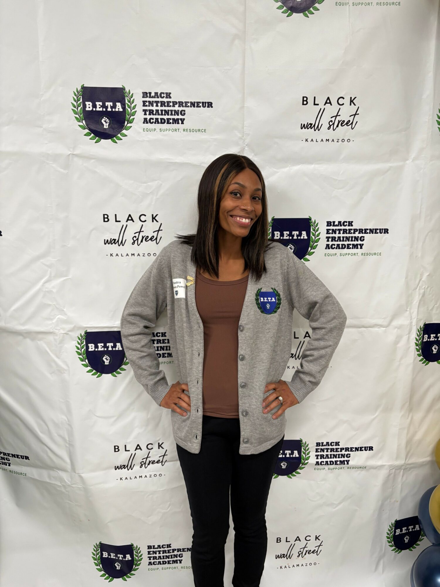 Woman standing in front of a white backdrop with logos, wearing a gray blazer, brown top, and black pants.
