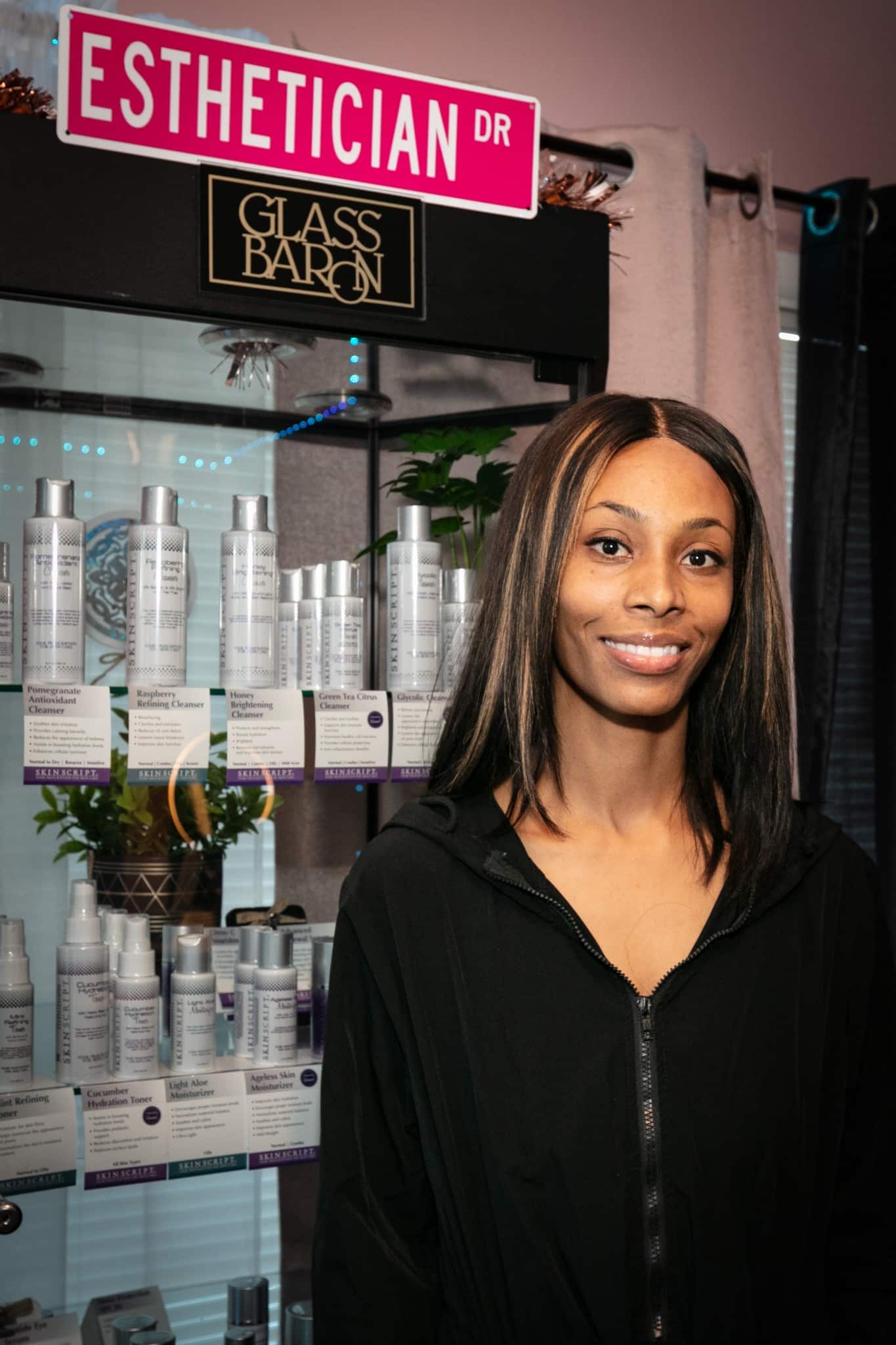 Smiling woman with shoulder-length hair in front of skincare products display, store interior, pink street sign, and black sign reading 'Glass Barn'.