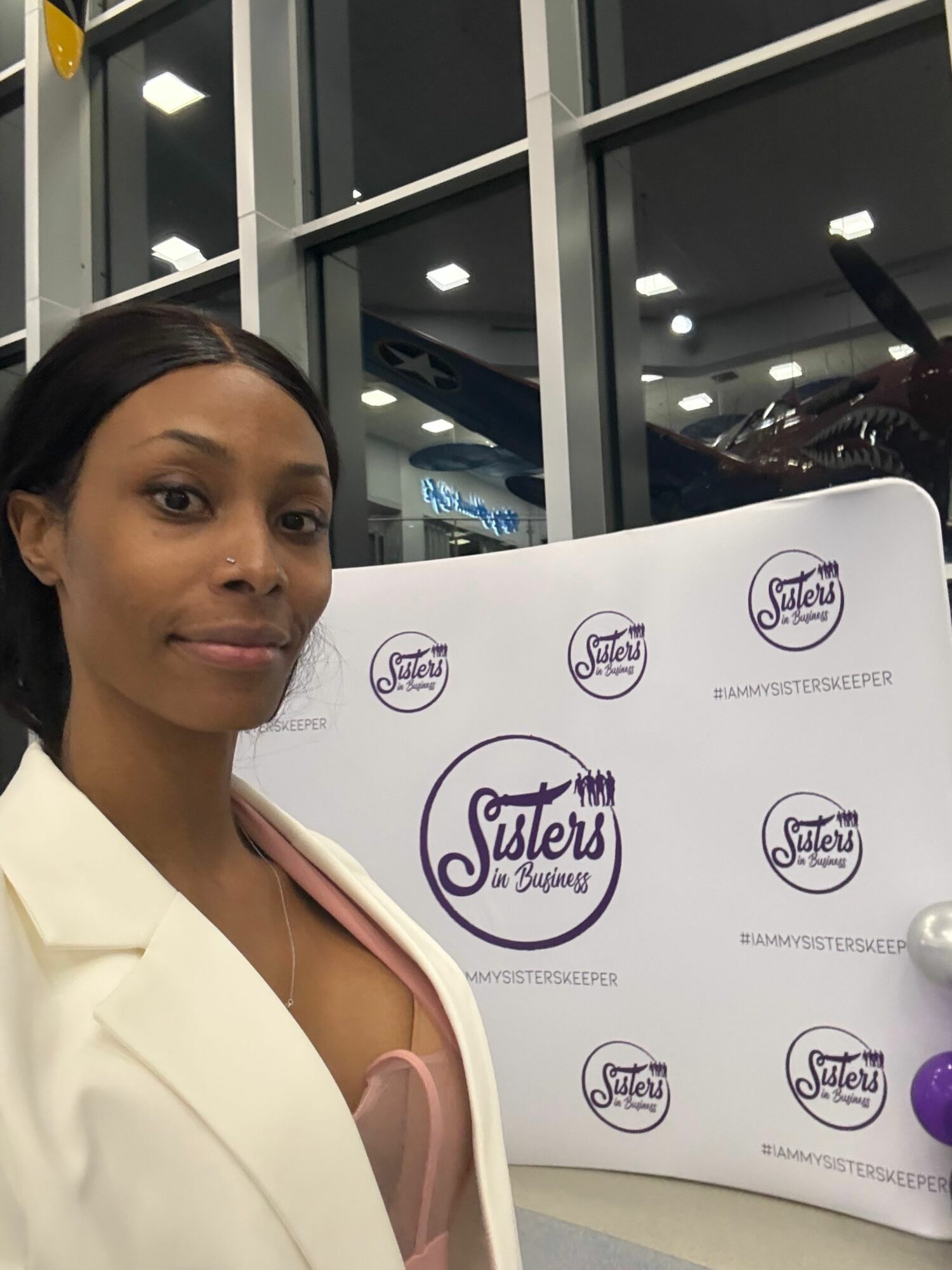 Woman with dark hair in a white blazer at a promotional event, standing in front of a branded backdrop.