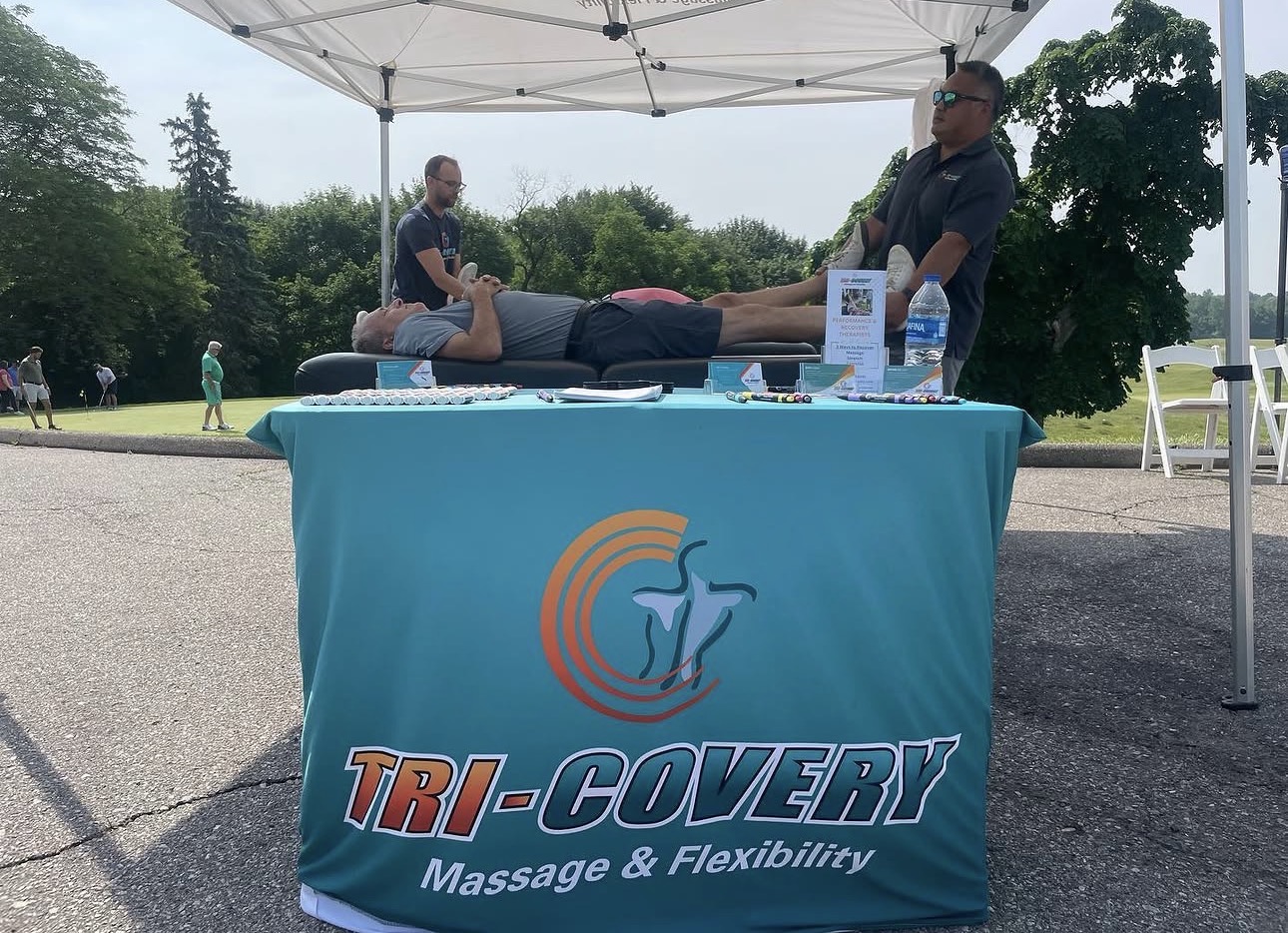 Person lying on a table under a canopy, receiving a massage outdoors, with two practitioners attending, and a table with a banner in front.