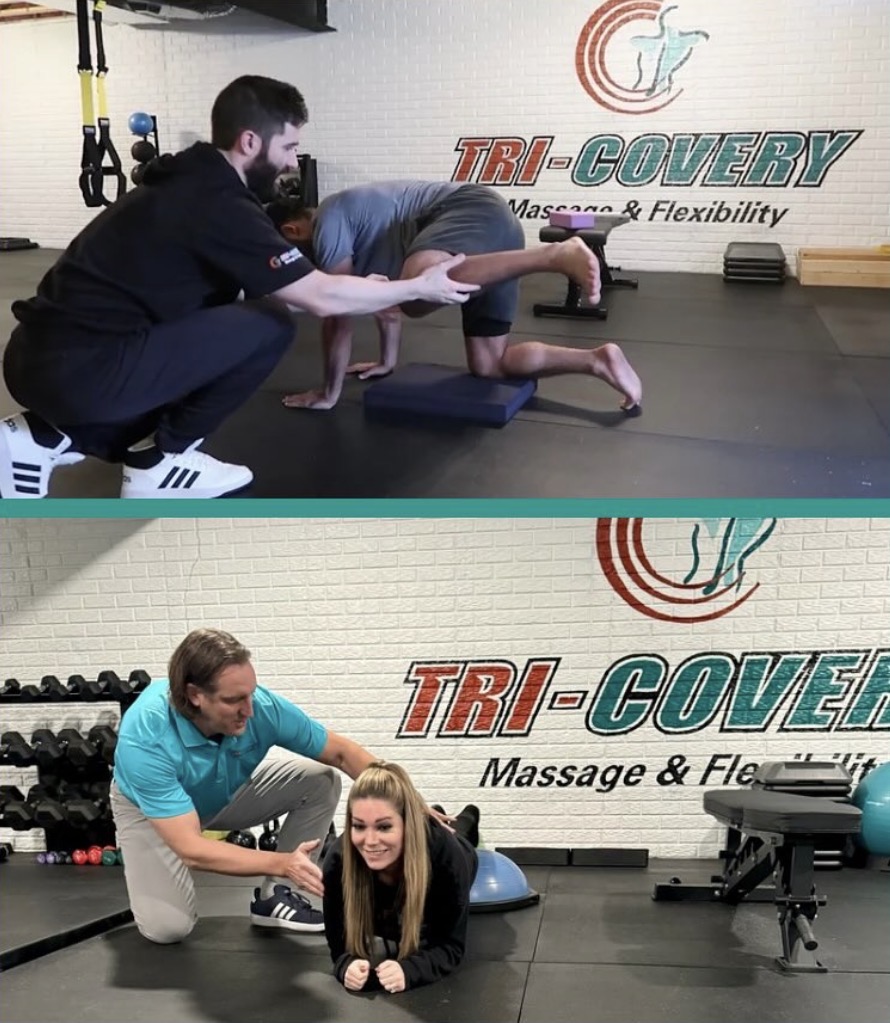 Trainer assisting a person in a plank position on a mat, with another person on hands and knees, in a gym. Logo and text on wall.
