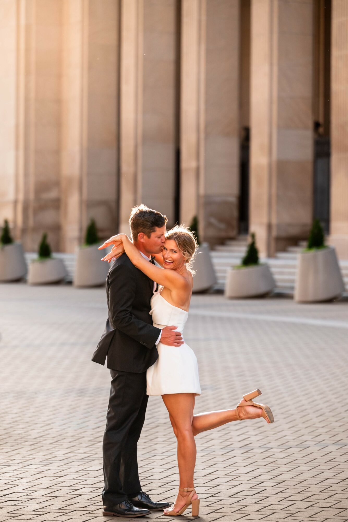A couple dancing outdoors in front of large columns, with the woman lifting her leg and smiling.