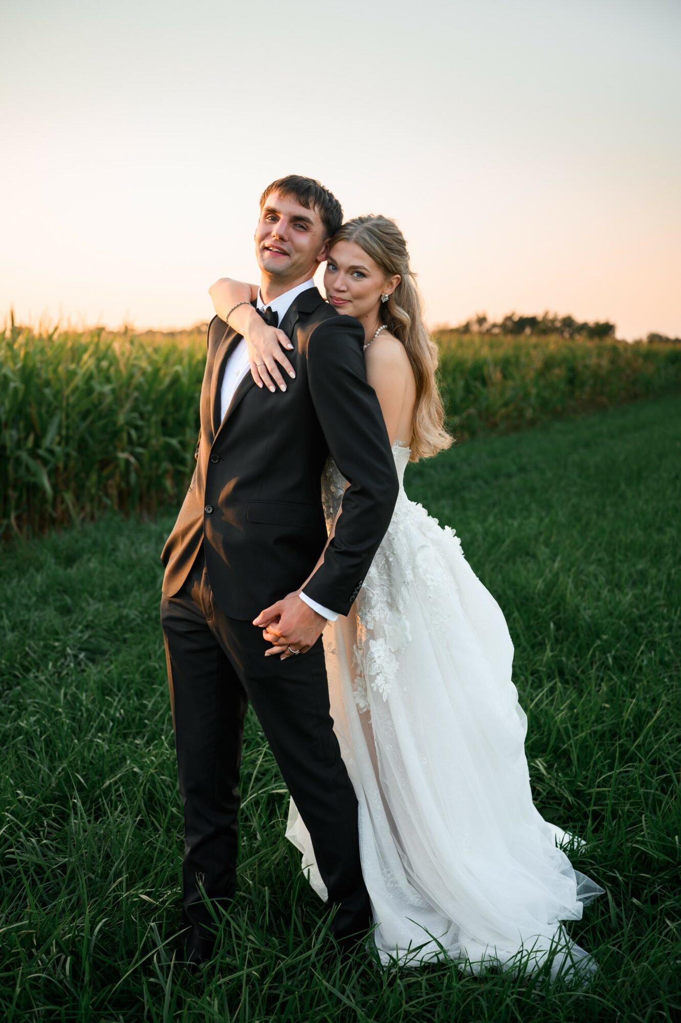 A bride and groom stand outdoors in a grassy field at sunset, smiling and embracing each other, with a cornfield in the background.