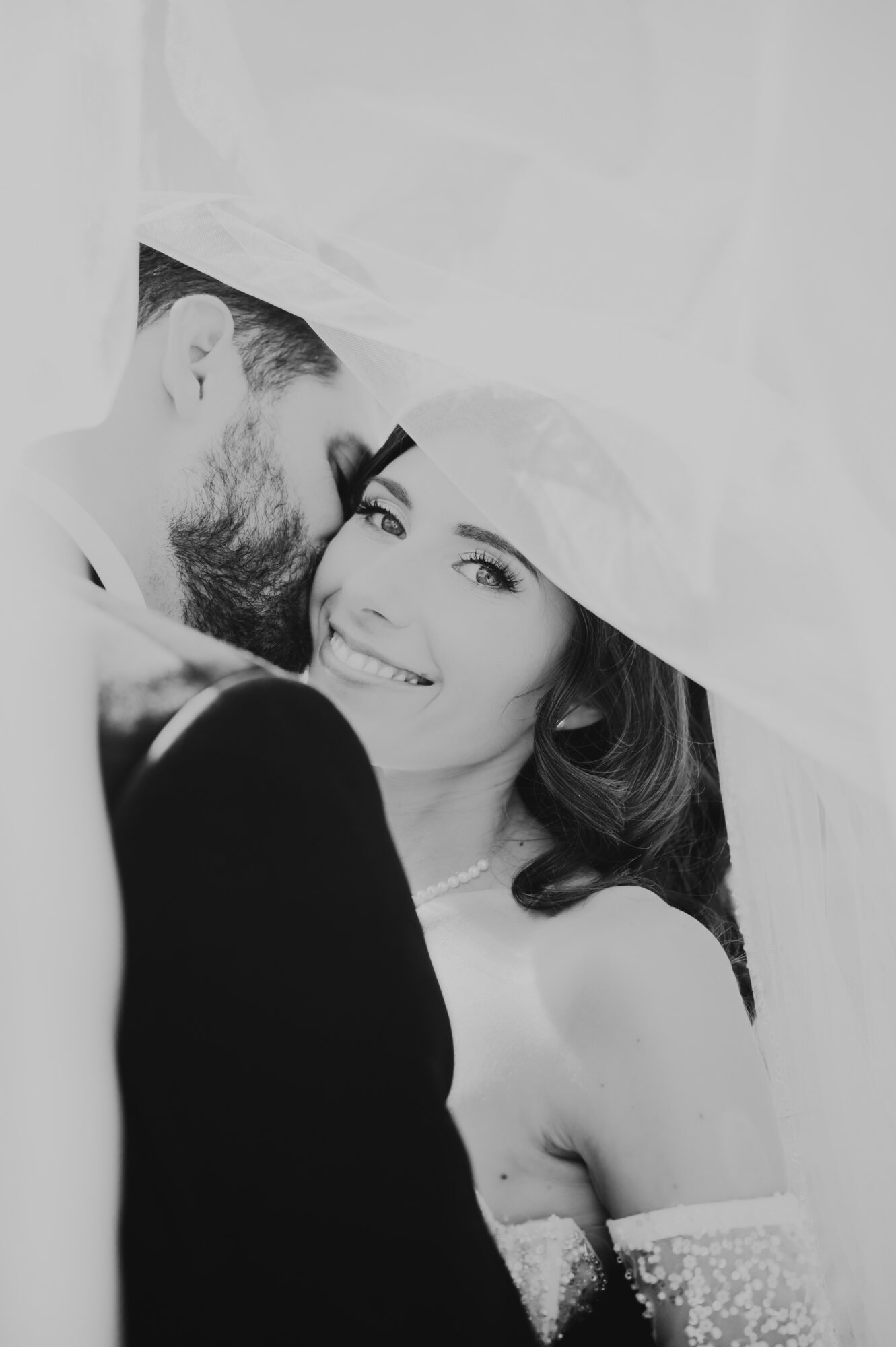 A smiling woman with dark hair and a man with a beard under a veil, close together, in black and white.