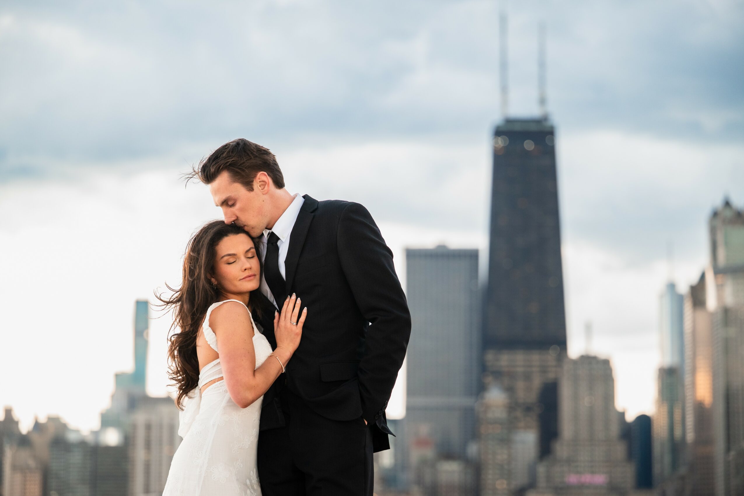 A man and woman embrace on a city rooftop with tall buildings and a cloudy sky in the background.