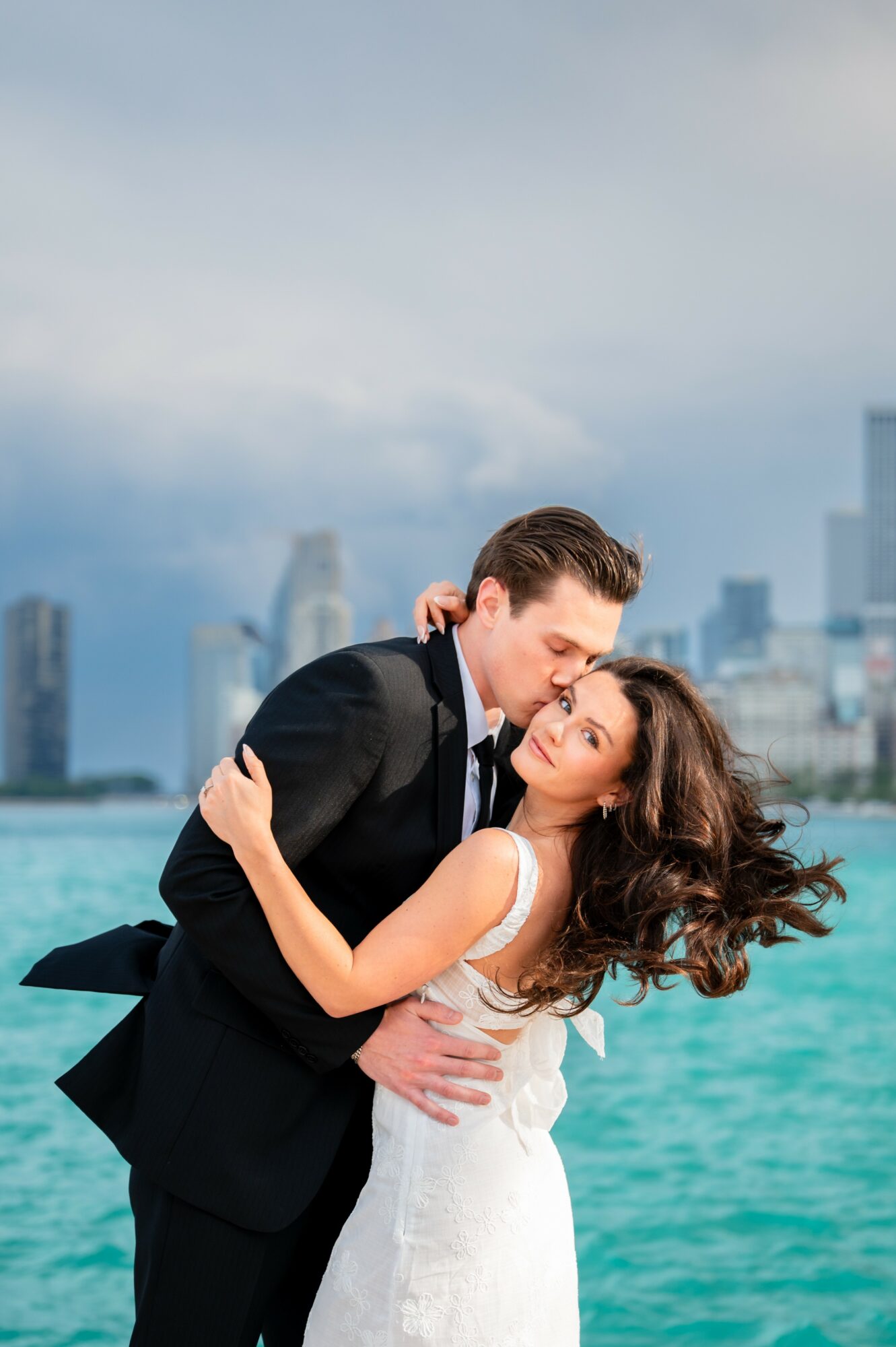 A couple in formal attire embracing on a waterfront with city skyline in background.