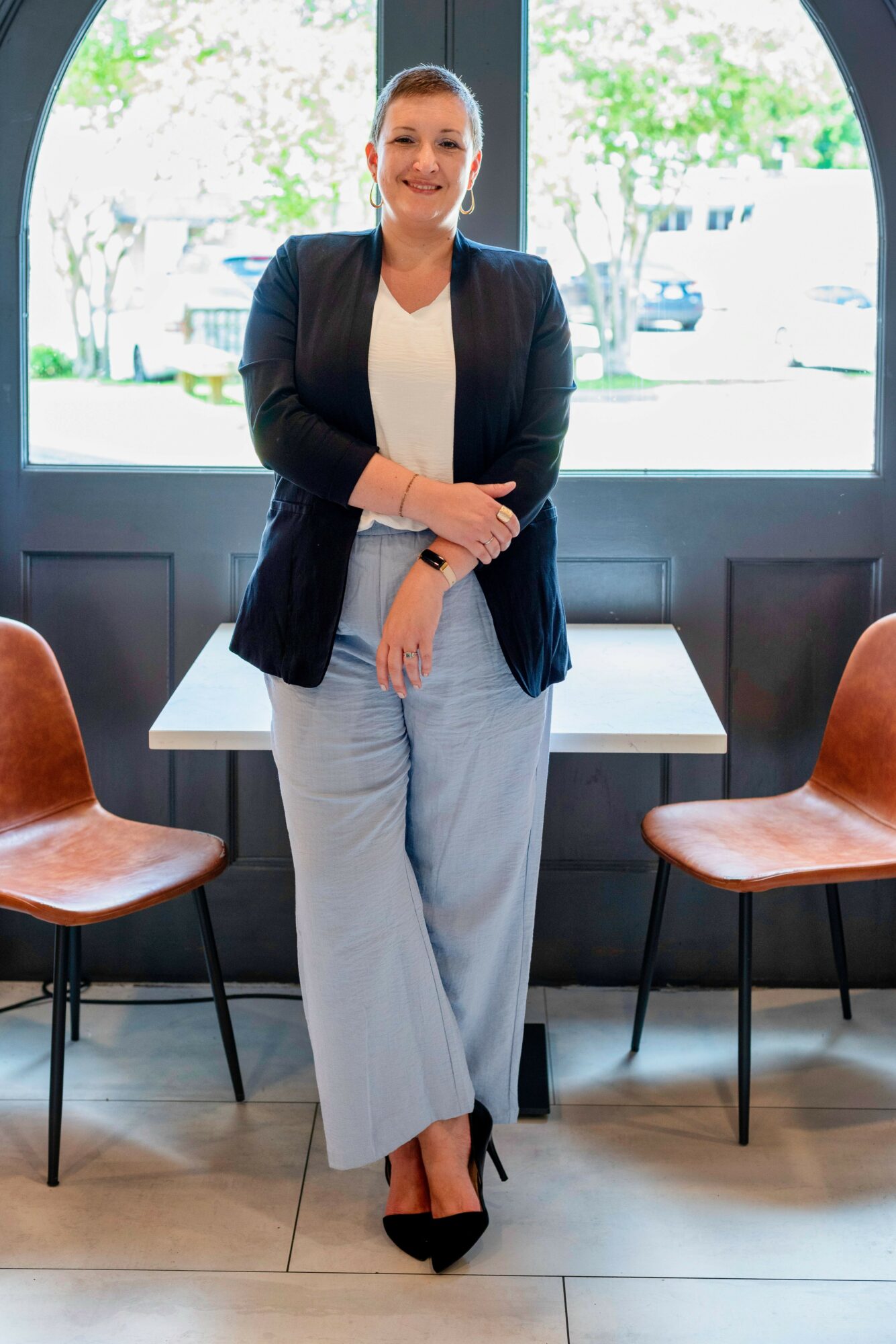 Woman standing indoors in front of a window, smiling, wearing a blazer, white top, and light-colored pants.