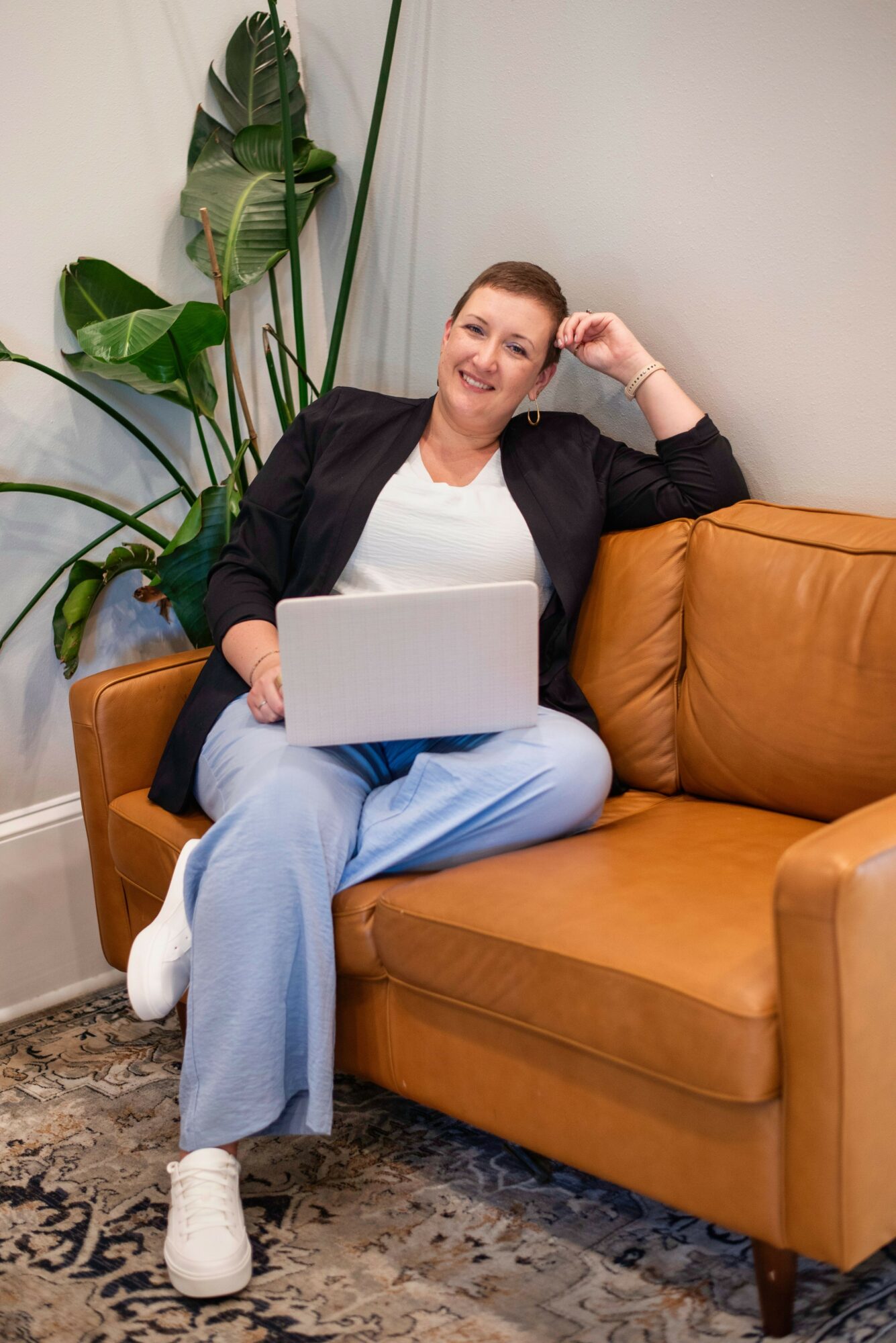 Person sitting on a brown sofa with a laptop, smiling, in a room with a large plant and a light-colored wall.