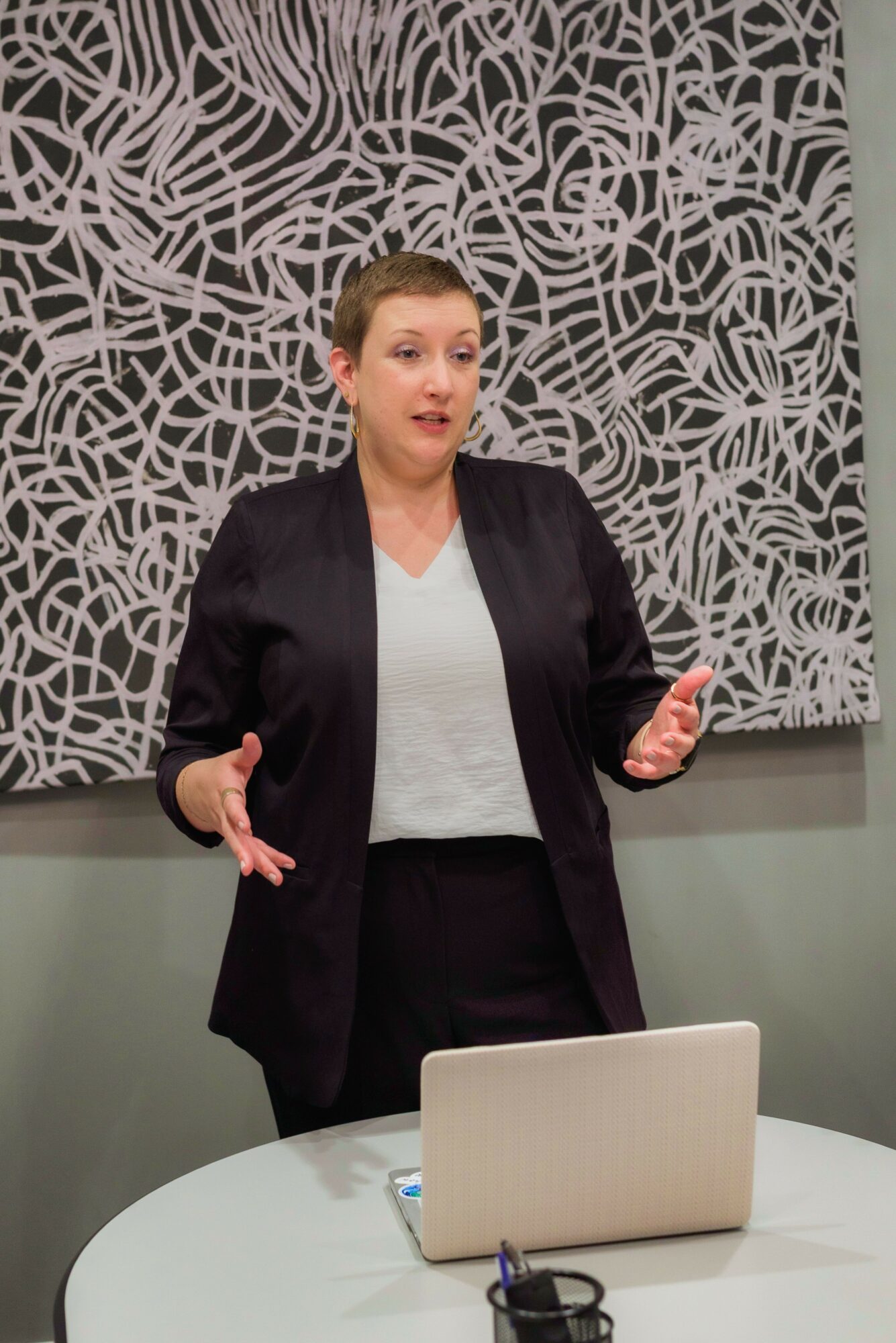 Woman speaking in front of patterned wall, gesturing with hands, standing behind a table with a laptop and cup.